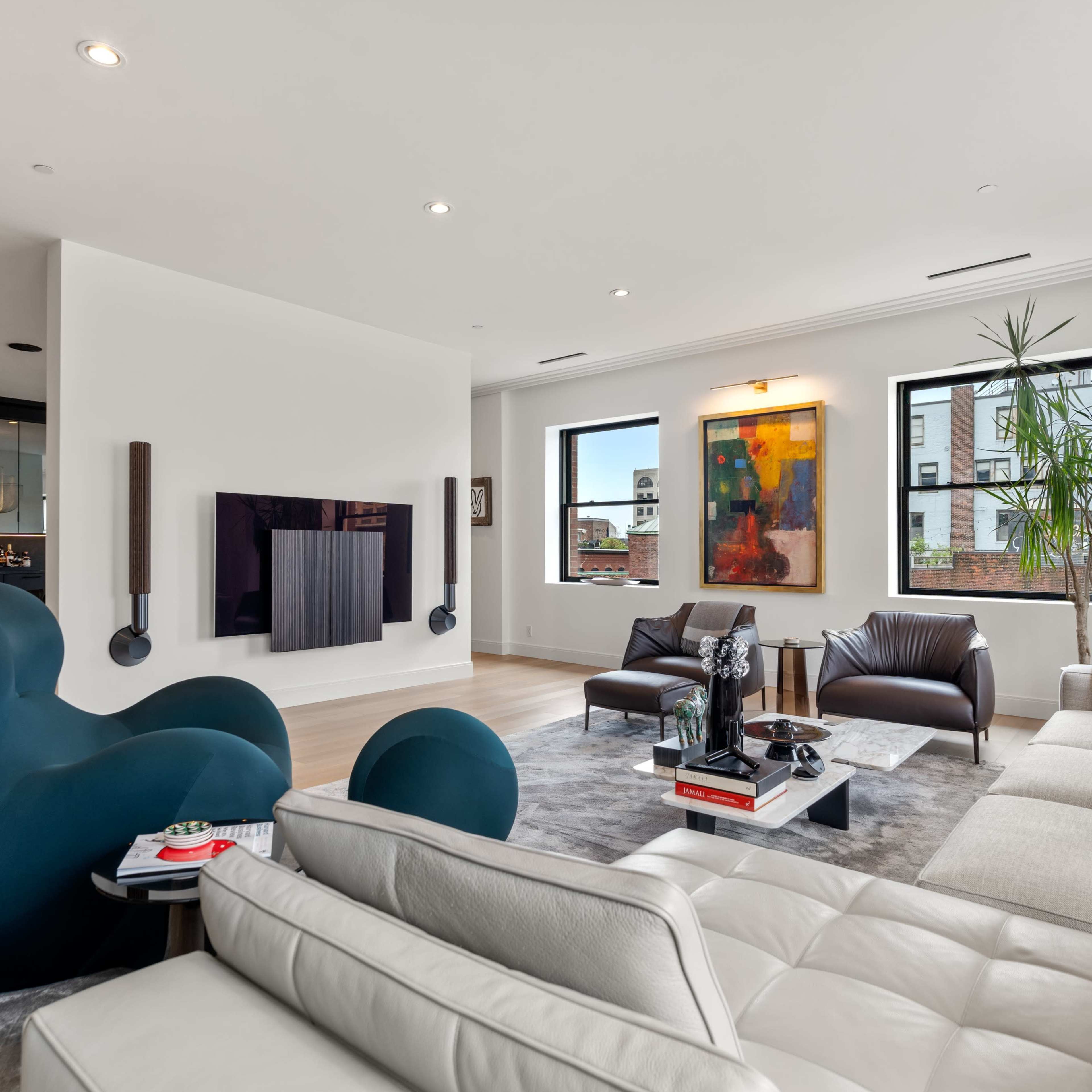 A modern living room features a mix of seating with a curved blue chair, a white leather sofa, and brown leather chairs, along with a large television mounted on the wall and vibrant artwork.