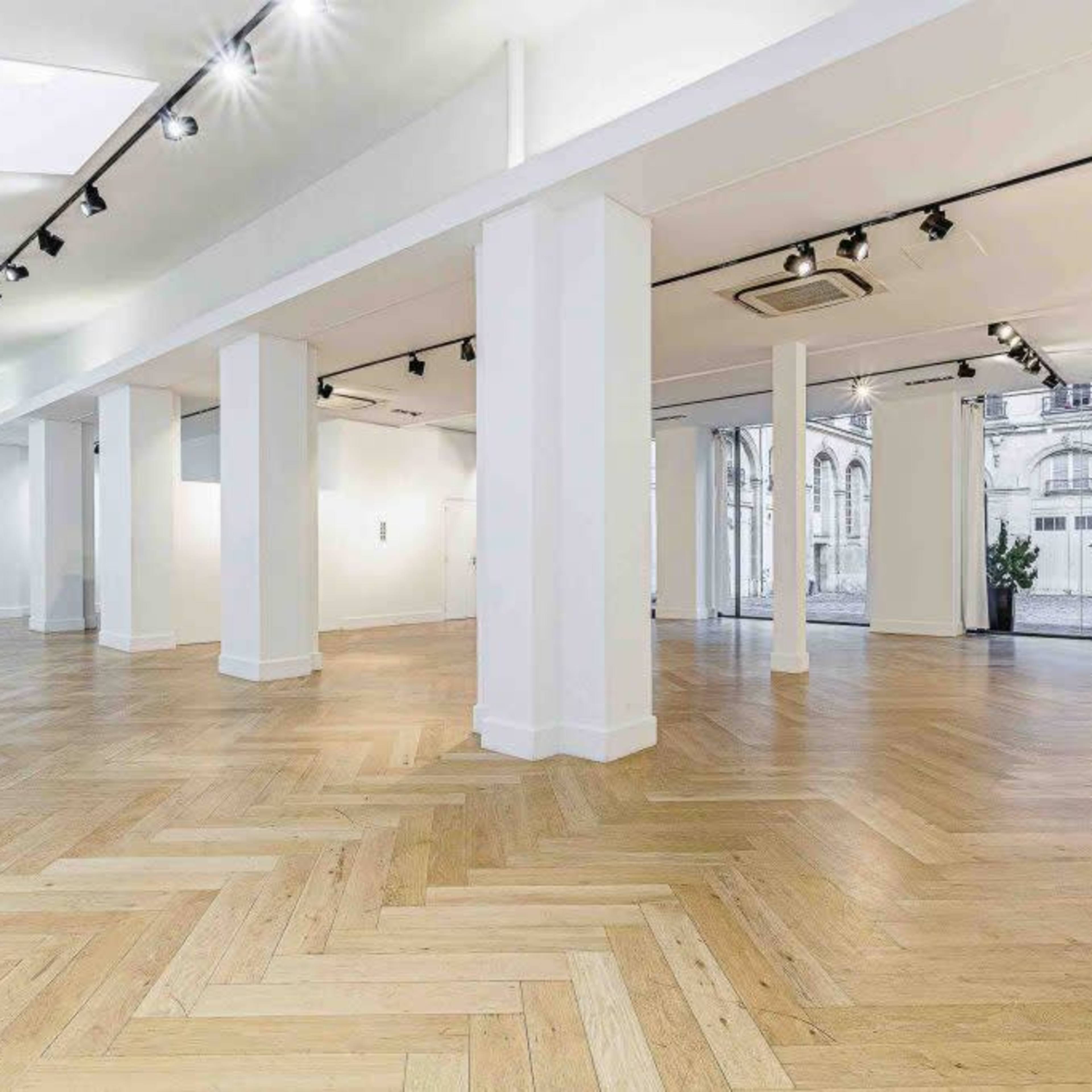 A spacious, empty gallery with a herringbone wood floor, white walls, and large windows.