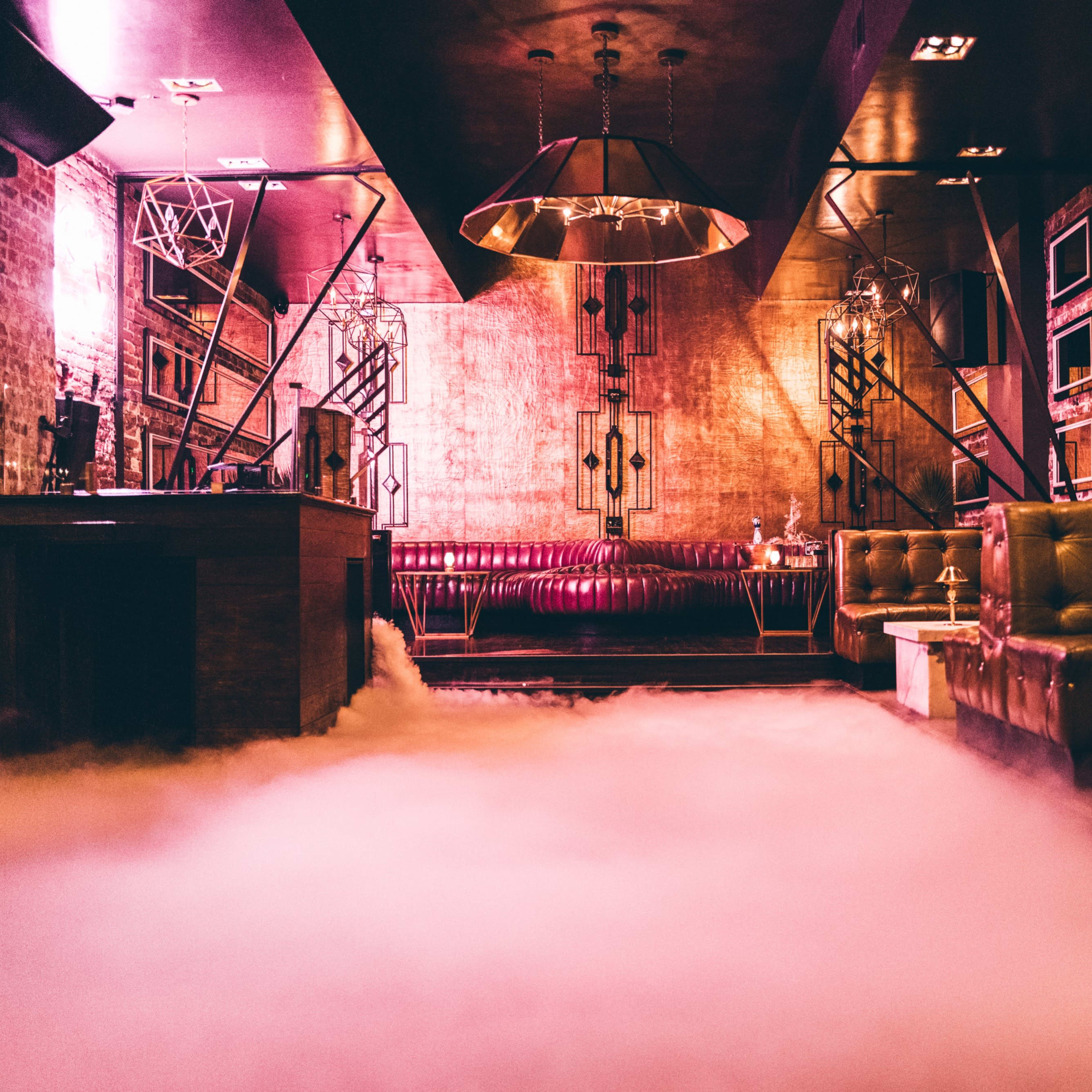 The interior of a dimly lit lounge features plush seating, a bar with marble-topped tables, and a fog effect covering the floor.