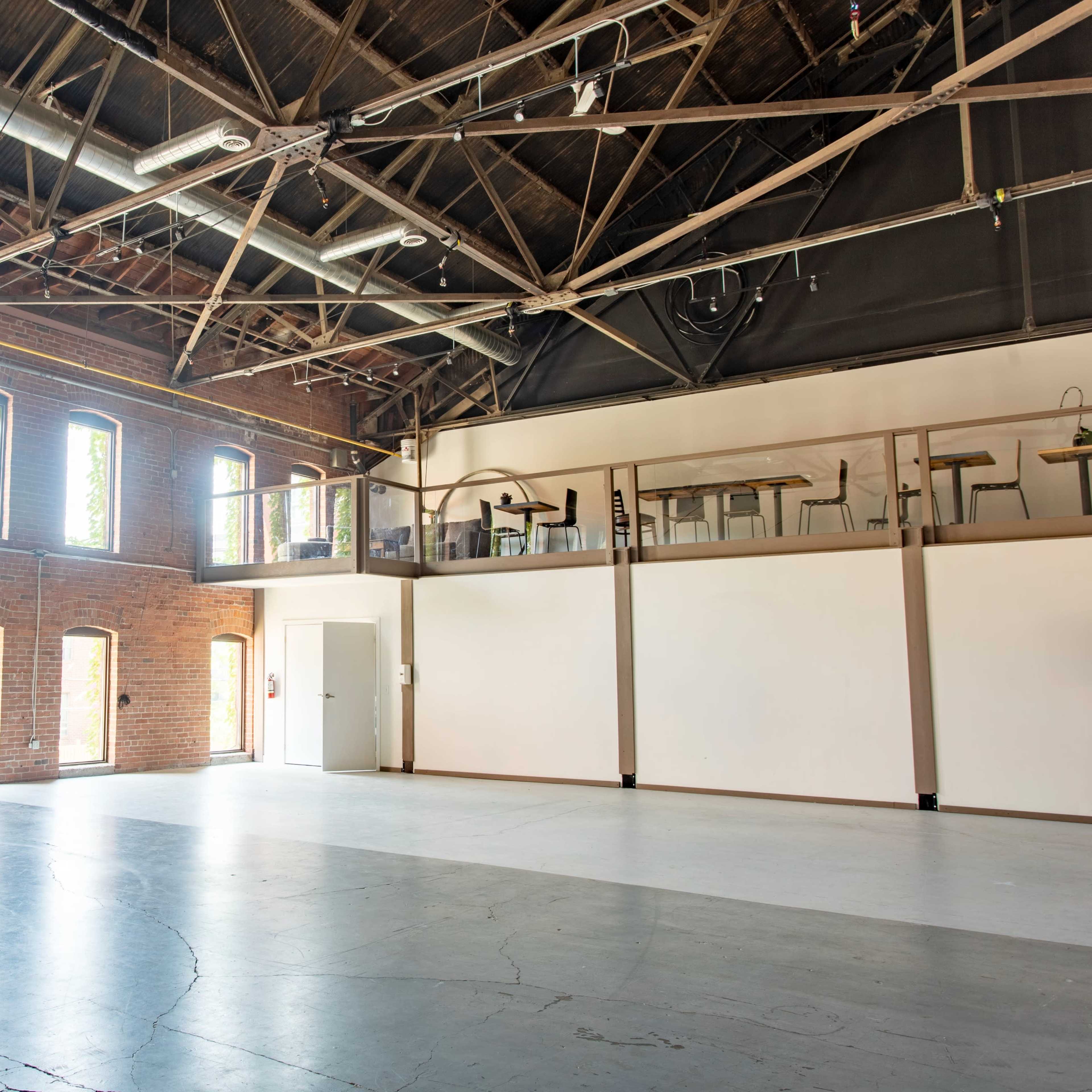An empty industrial space with tall brick walls, large windows, and a lofted area overlooking the main floor.