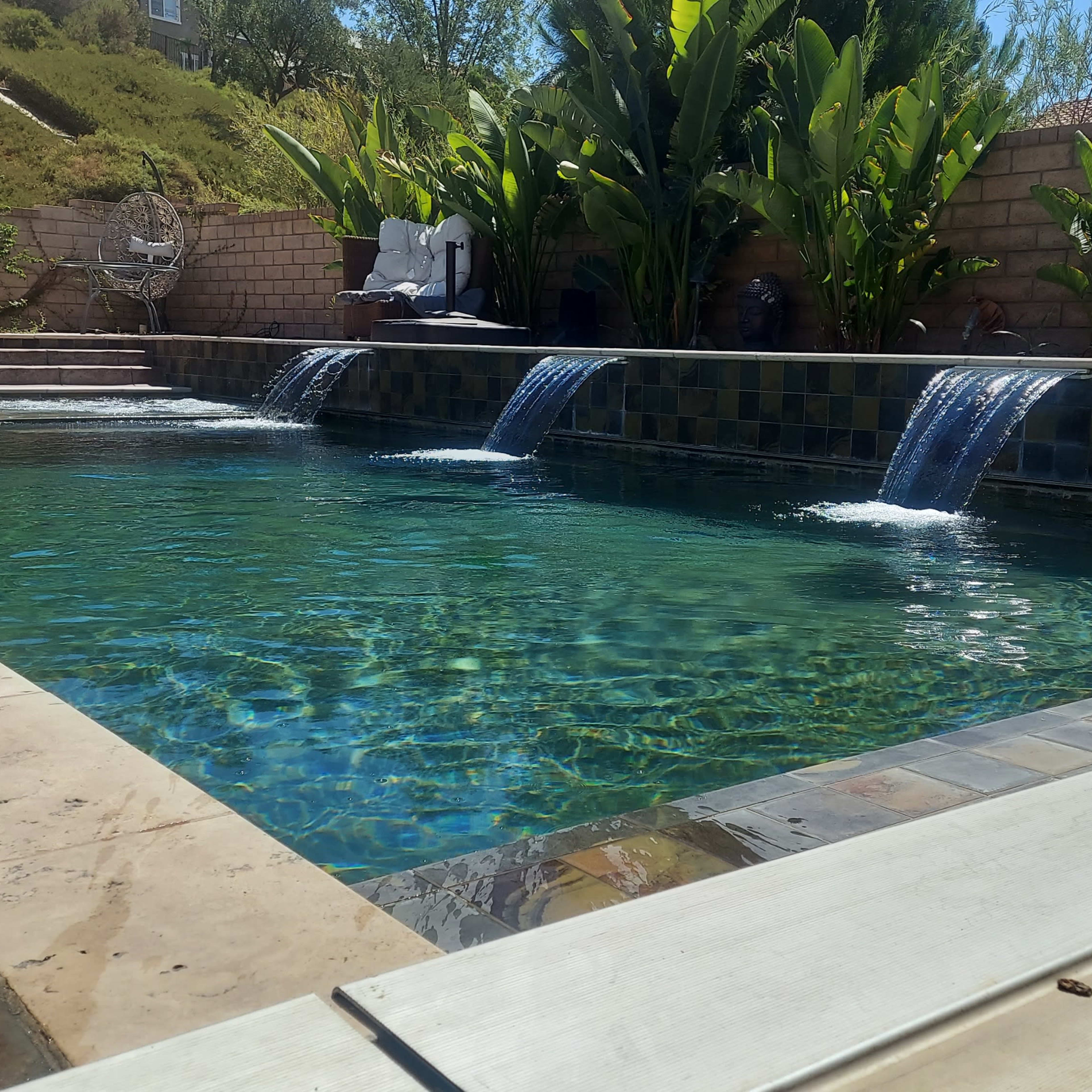 A swimming pool with three water features spilling into it, surrounded by lush greenery and patio furniture.