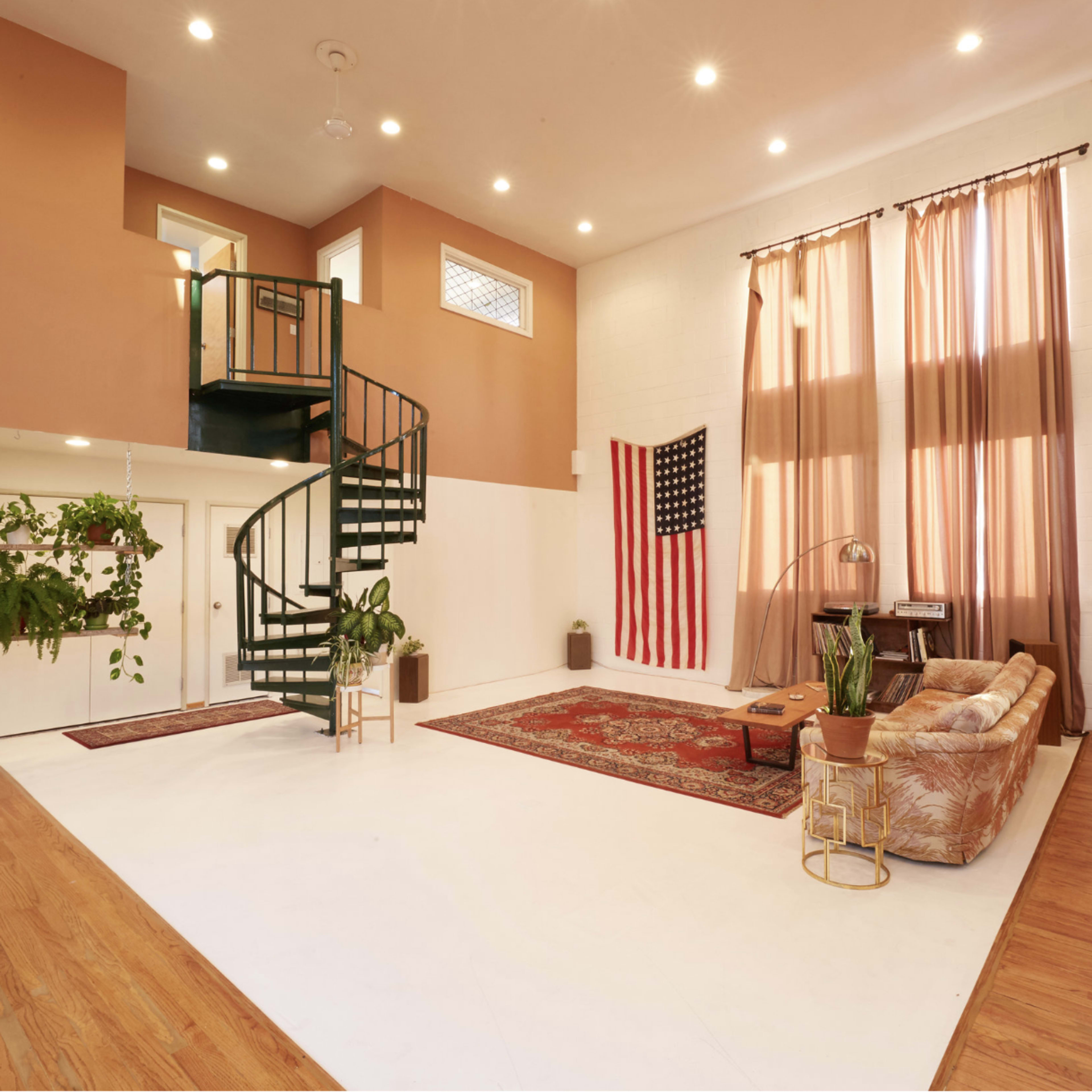 A spacious living area features a spiral staircase, large windows with draped curtains, an American flag, potted plants, and a mix of furniture on hardwood floors.