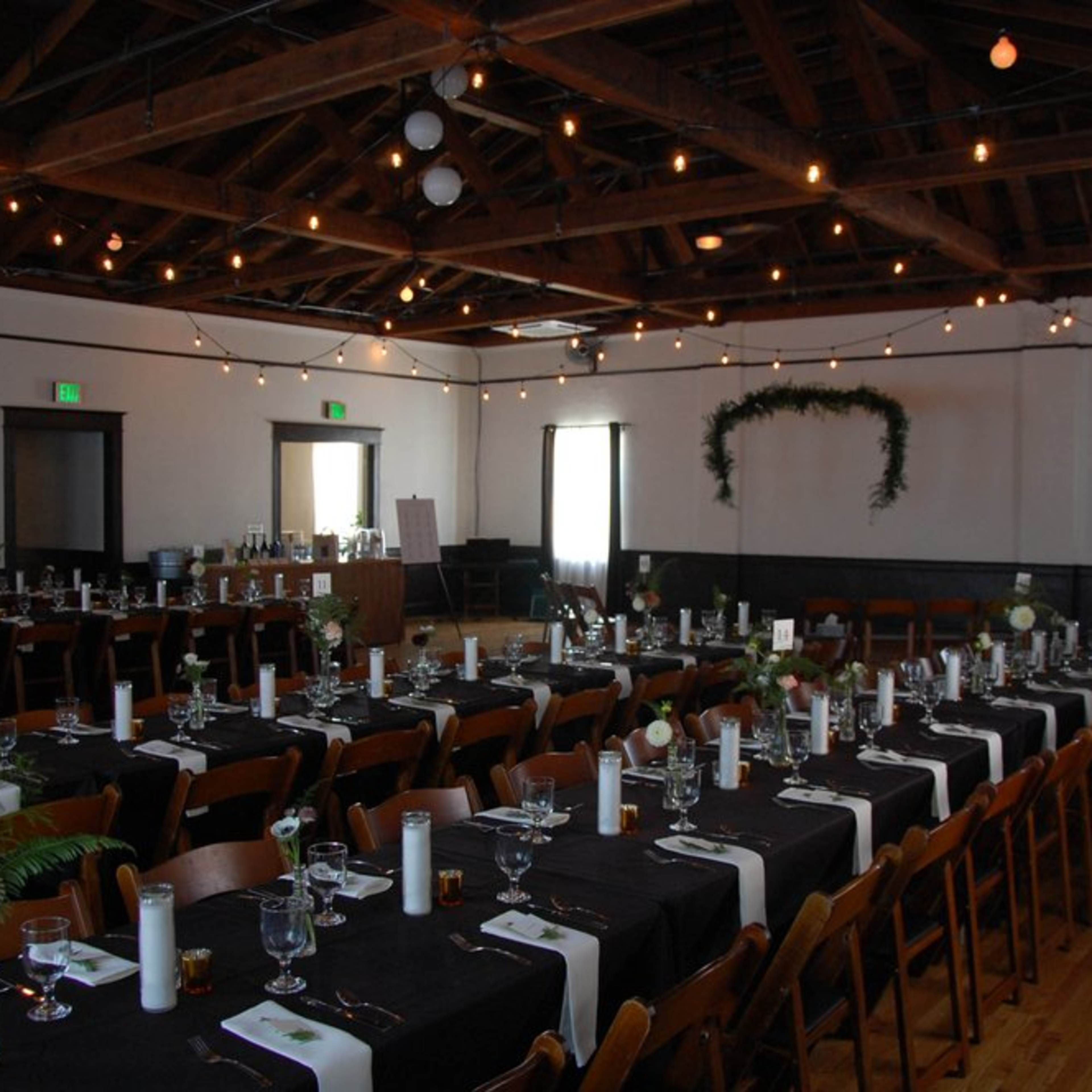 A large dining area with long tables arranged for an event, decorated with black tablecloths, white candles, and a greenery backdrop.