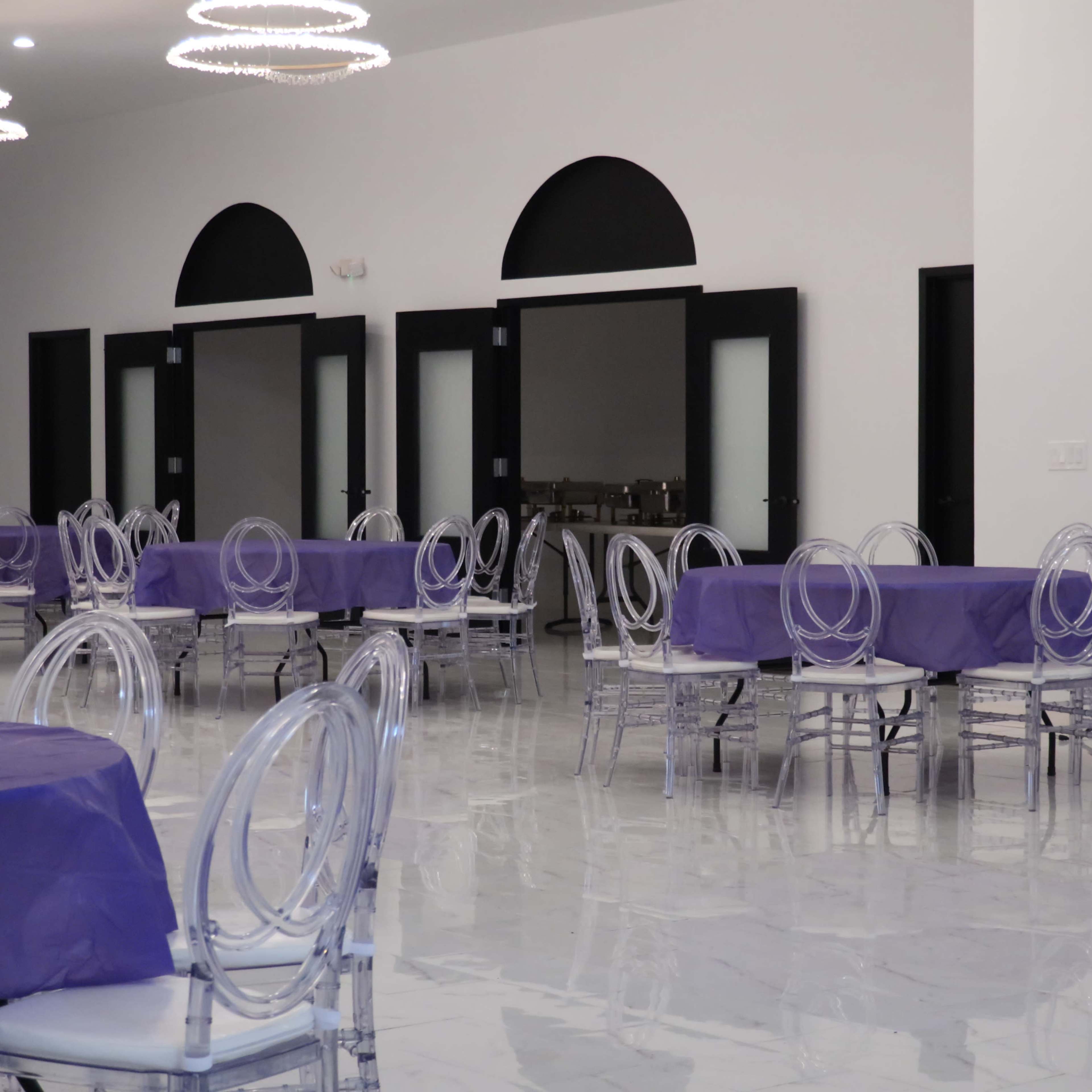 A spacious event room with several round tables covered in purple tablecloths and clear acrylic chairs, alongside large black-framed doors.