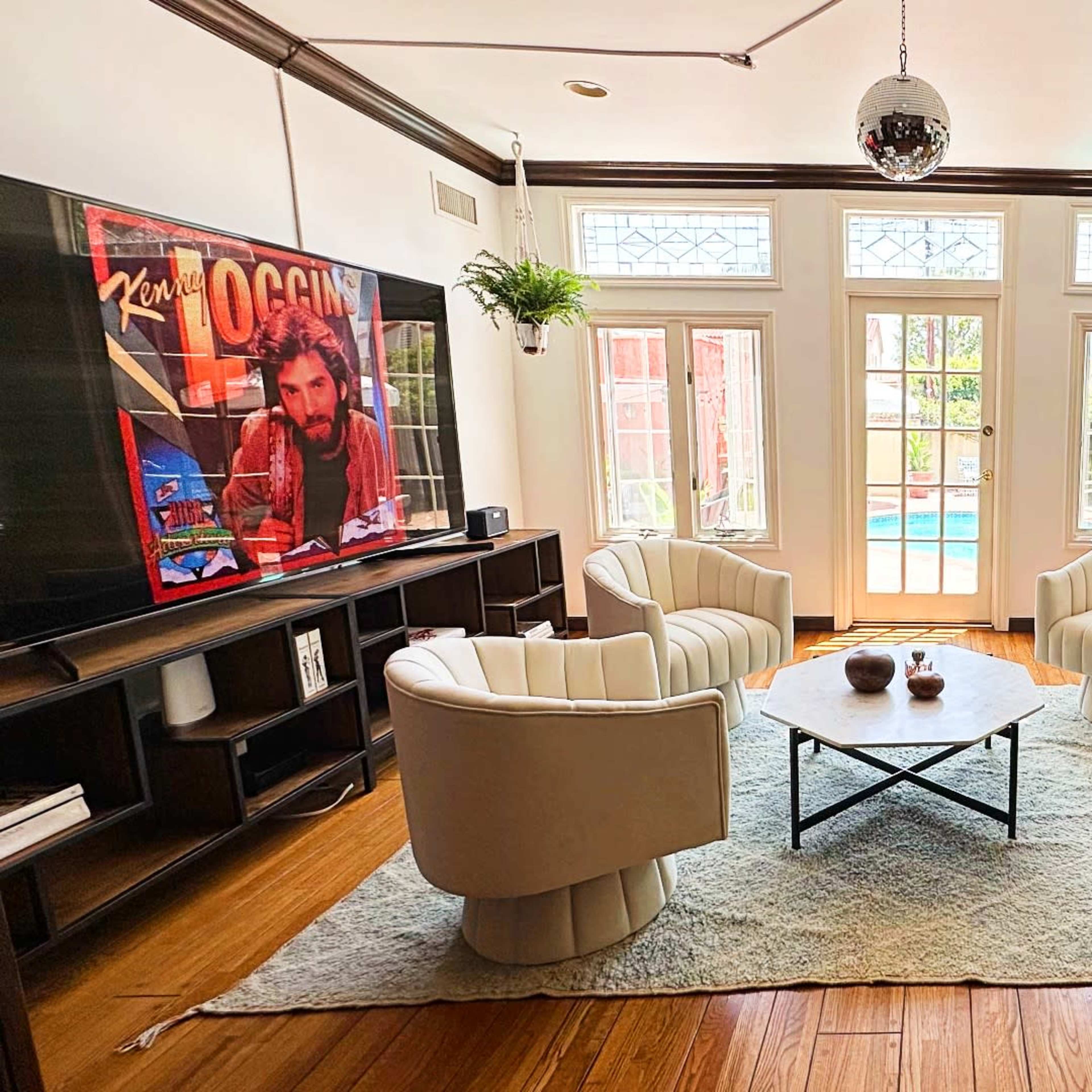 A living room featuring four white chairs arranged around a black coffee table on a light rug, with a large television mounted on the wall and doors leading to a pool area in the background.