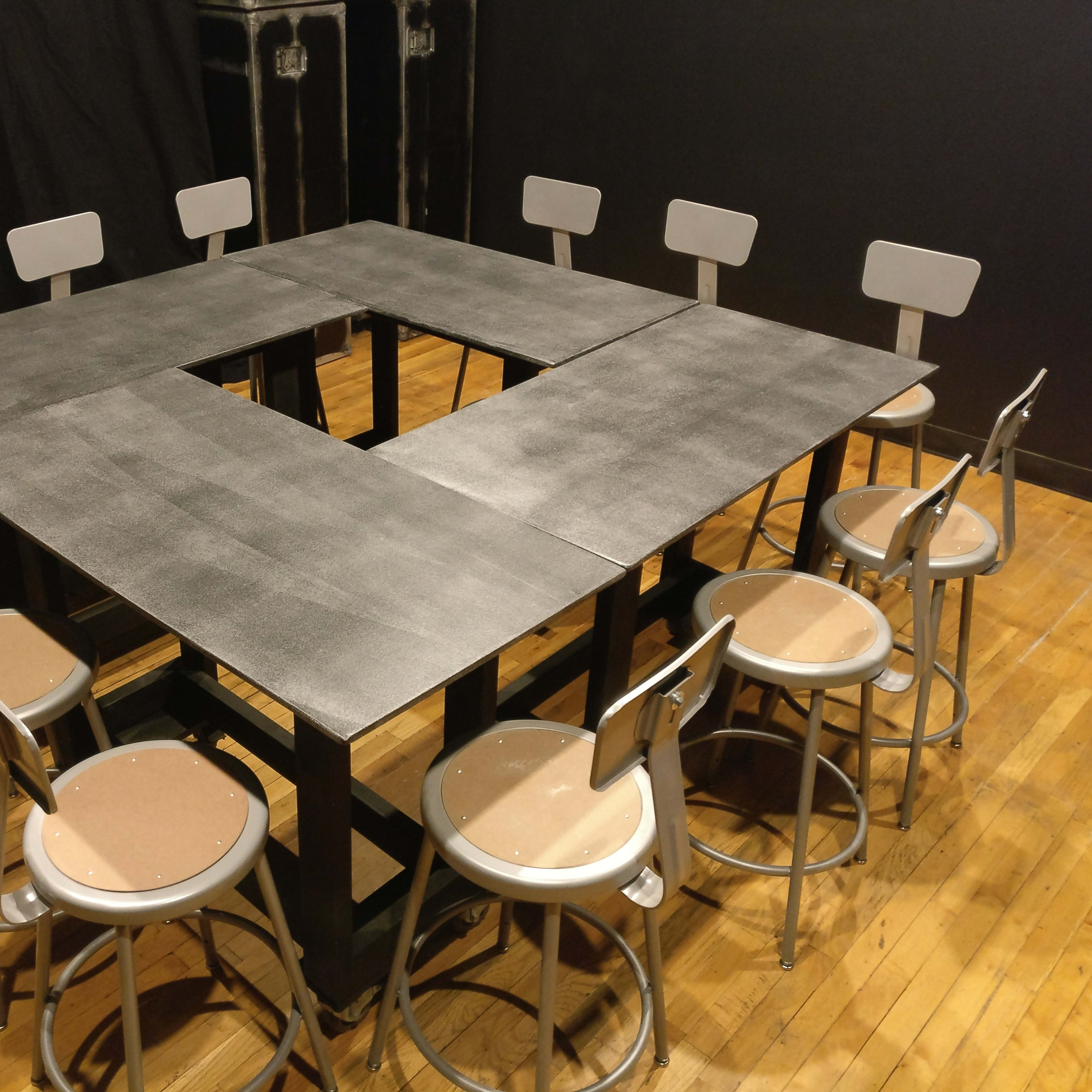 A square table with a cutout in the center is surrounded by eight metal stools on a wooden floor.