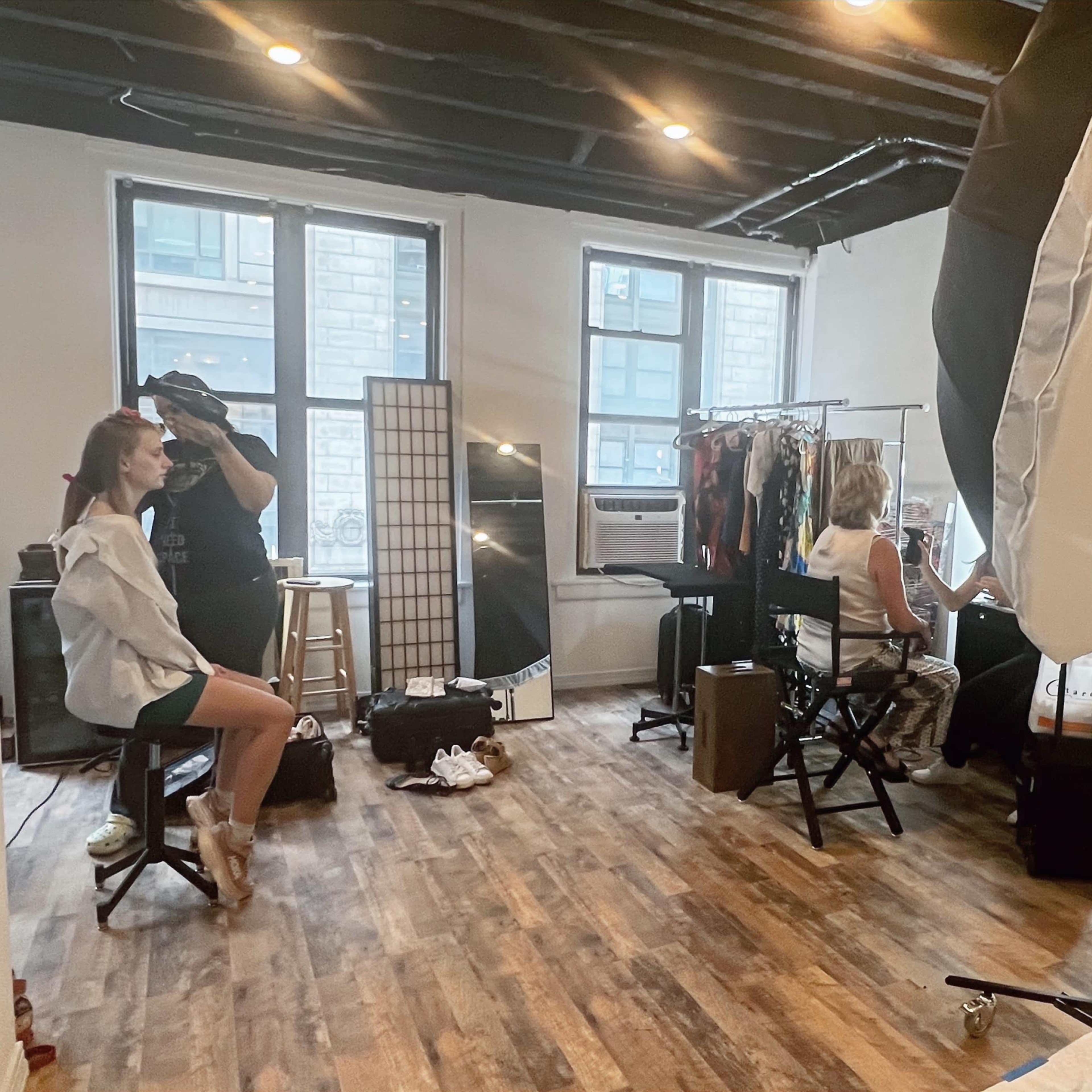 A model sits on a stool in a brightly lit room while a photographer adjusts equipment and a makeup artist prepares at a nearby station.