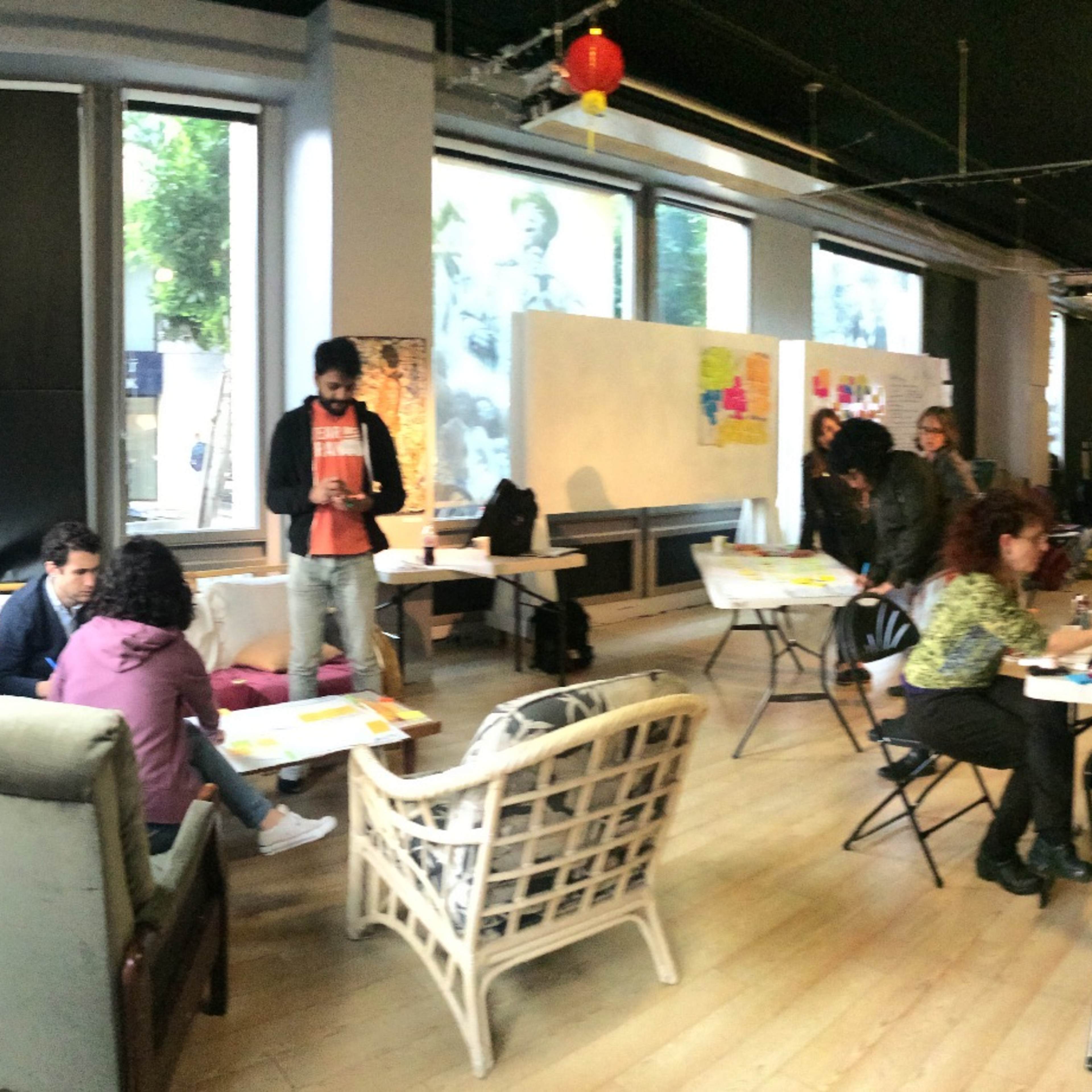 A panoramic view of a workspace featuring several tables where people are engaged in discussions and activities, with boards and posters on the walls.