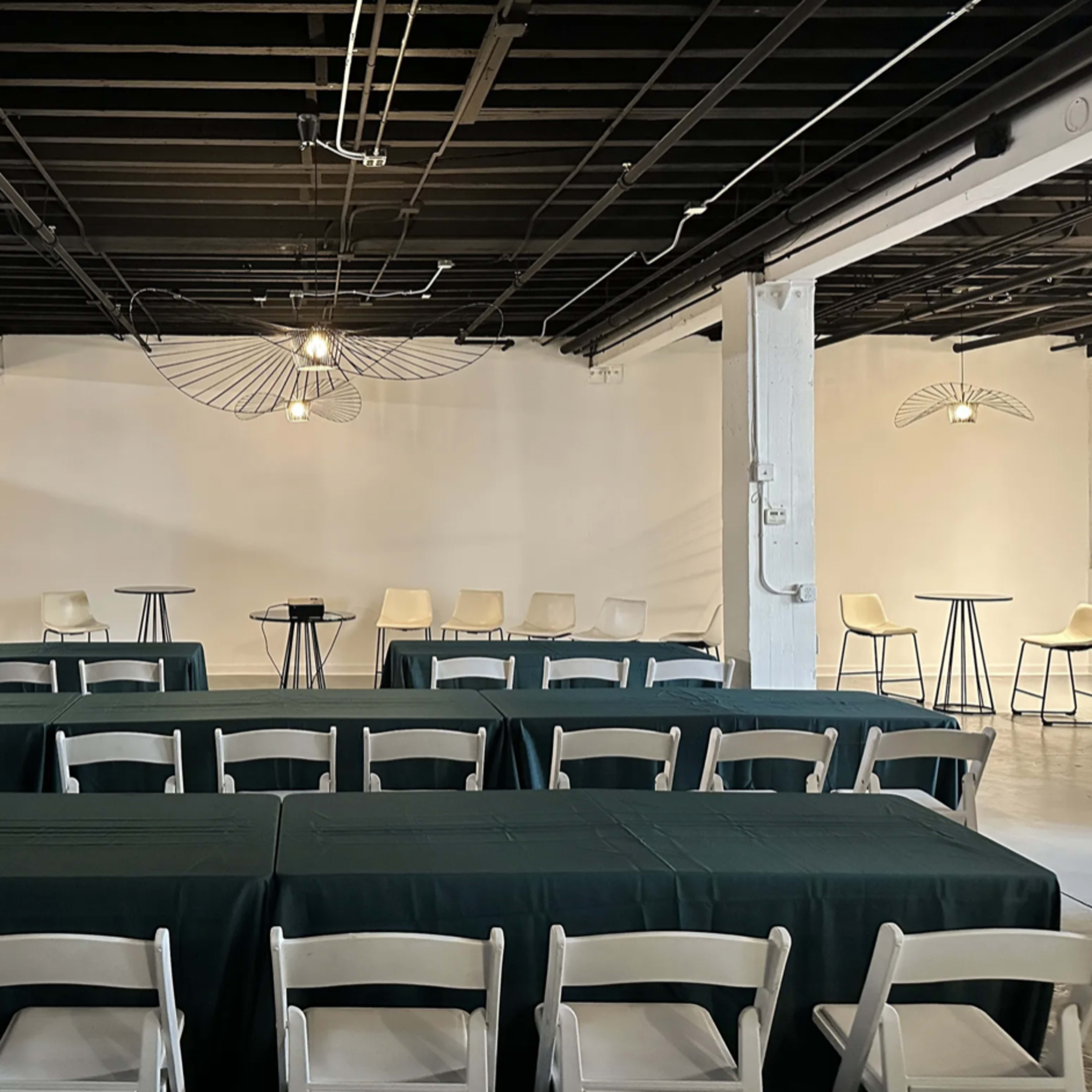 A spacious indoor venue arranged with green tablecloths covering long tables and a few high-top tables in a minimalist setting.