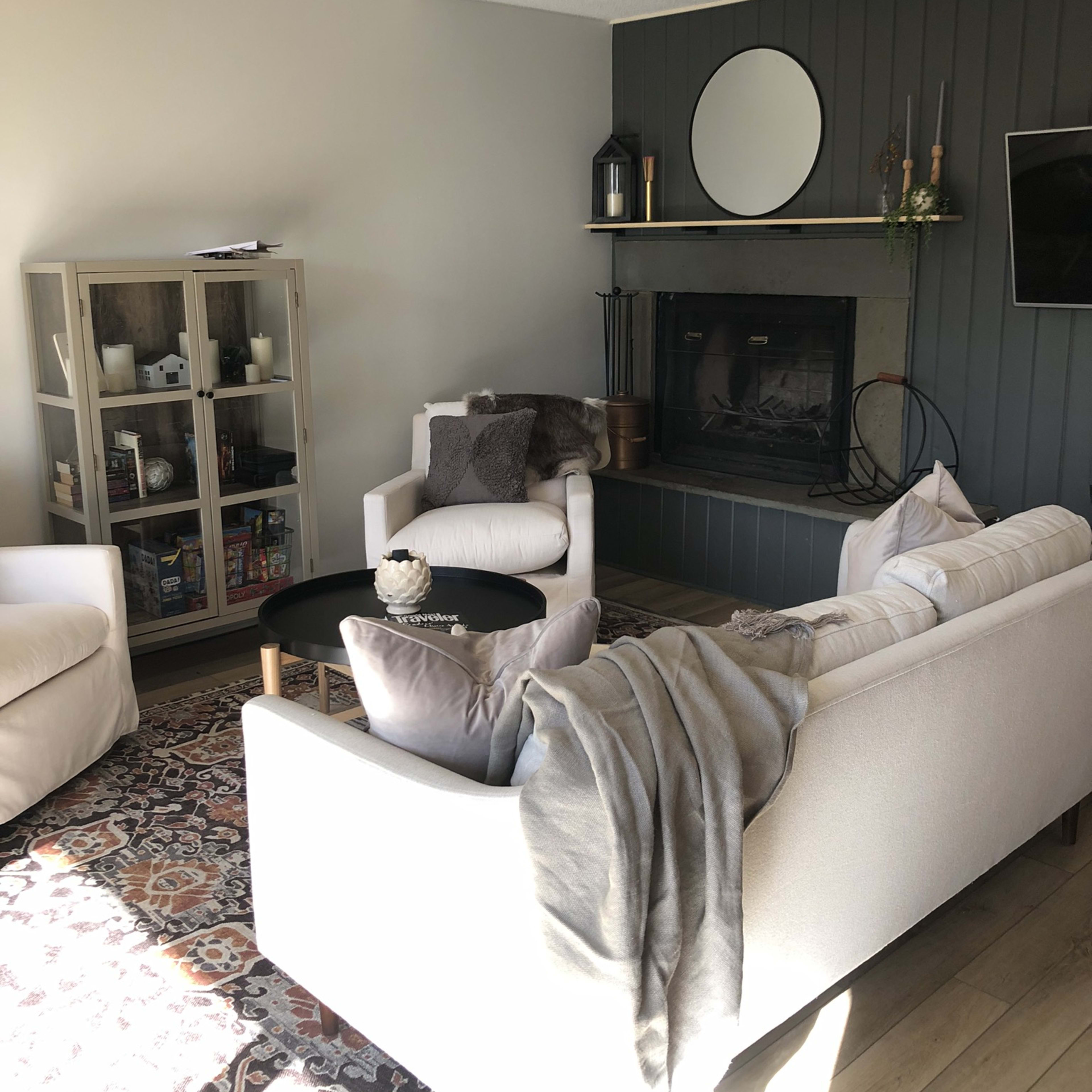 A cozy living room with a white sofa, two armchairs, a circular coffee table, and a fireplace, topped by a round mirror and decorated with plants.