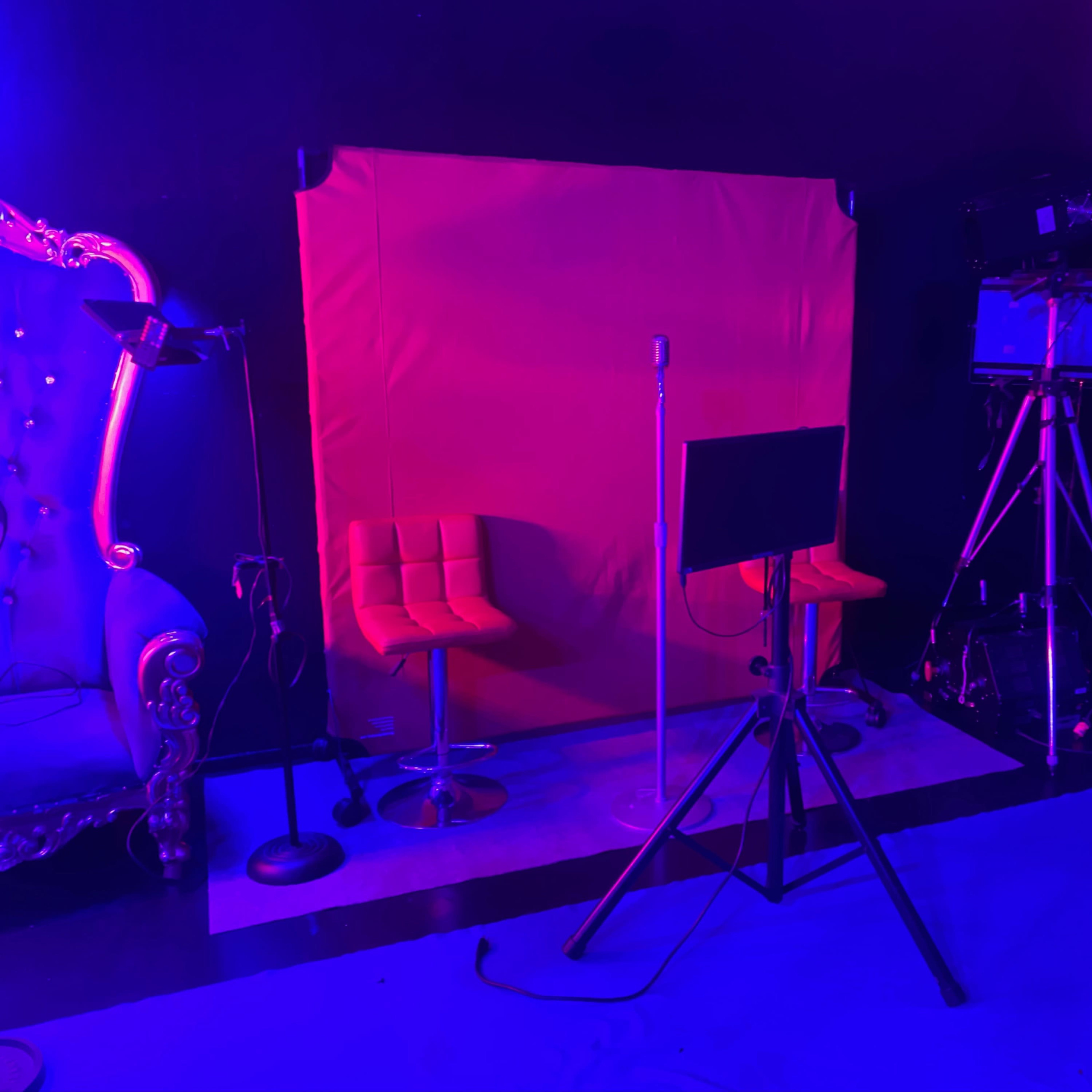 A studio setup with two orange chairs, a red backdrop, a microphone stand, and various lighting and filming equipment.