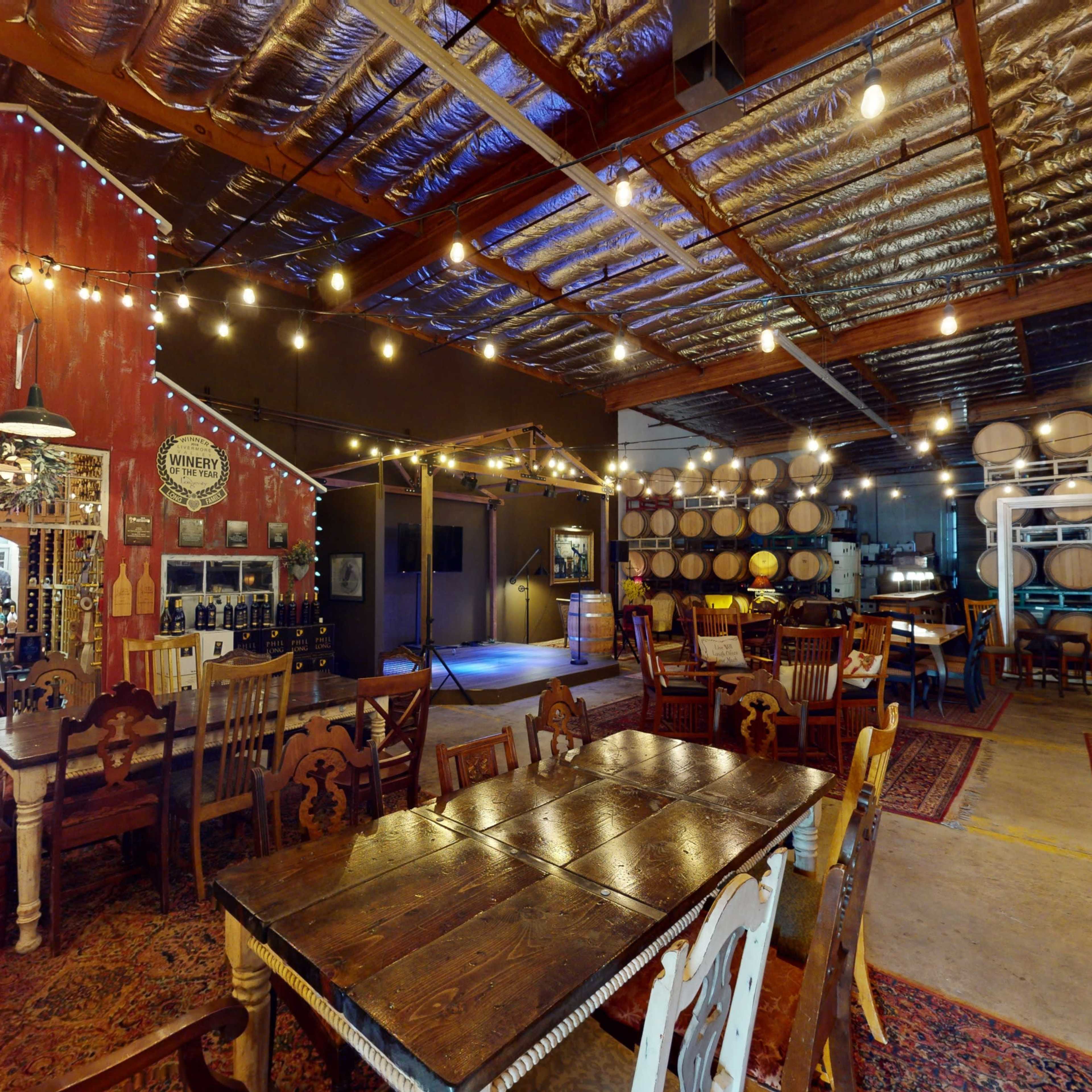 A rustic wine tasting room with wooden tables, a barn-style wall, and rows of wine barrels in the background, illuminated by string lights.