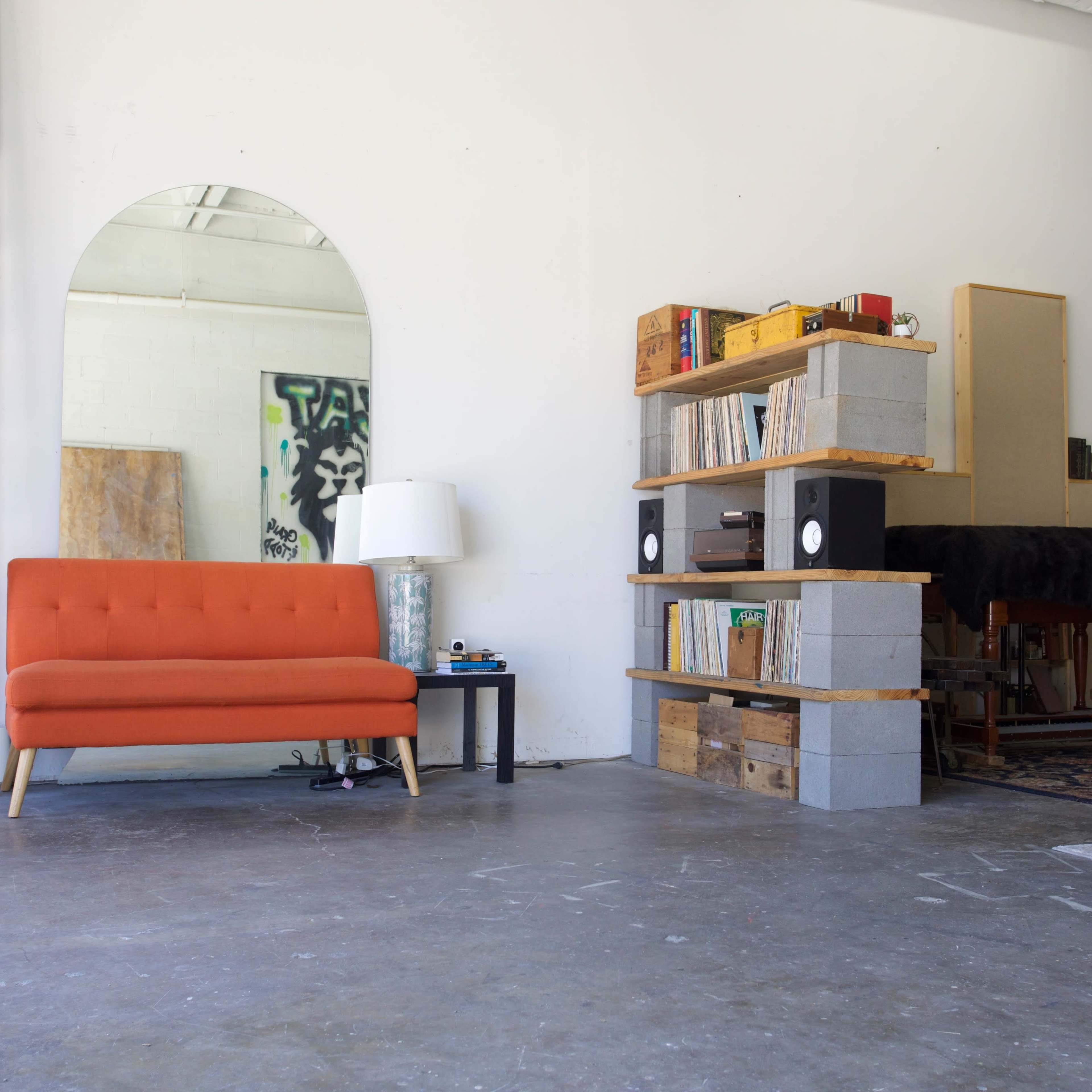 A spacious room featuring an orange sofa, a tall ladder, a large mirror, a bookshelf made of wooden crates and concrete blocks, and a table with speakers.