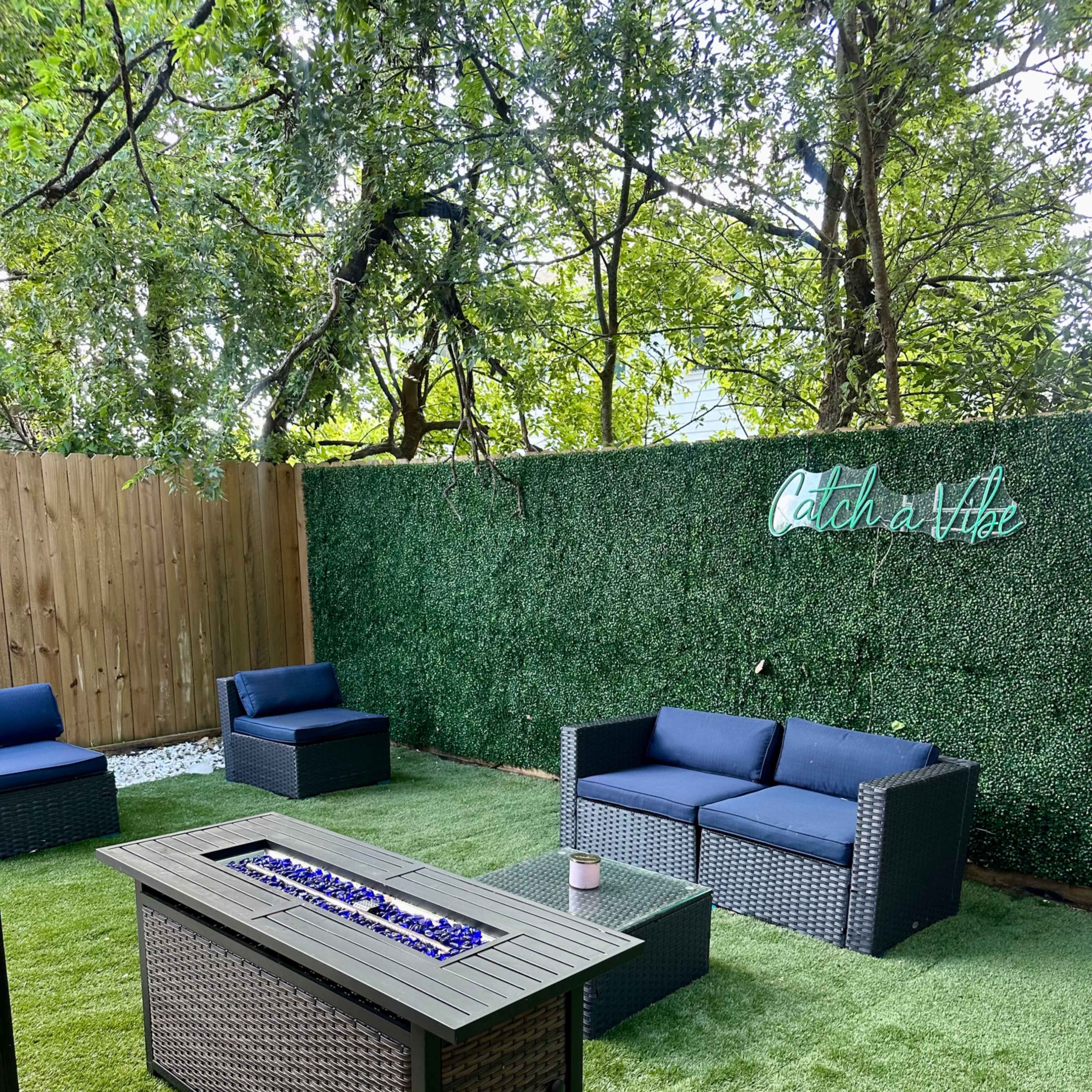 A landscaped outdoor seating area with two blue couches, a fire table with blue glass stones, and a green hedge backdrop featuring the phrase "Catch a Vibe."