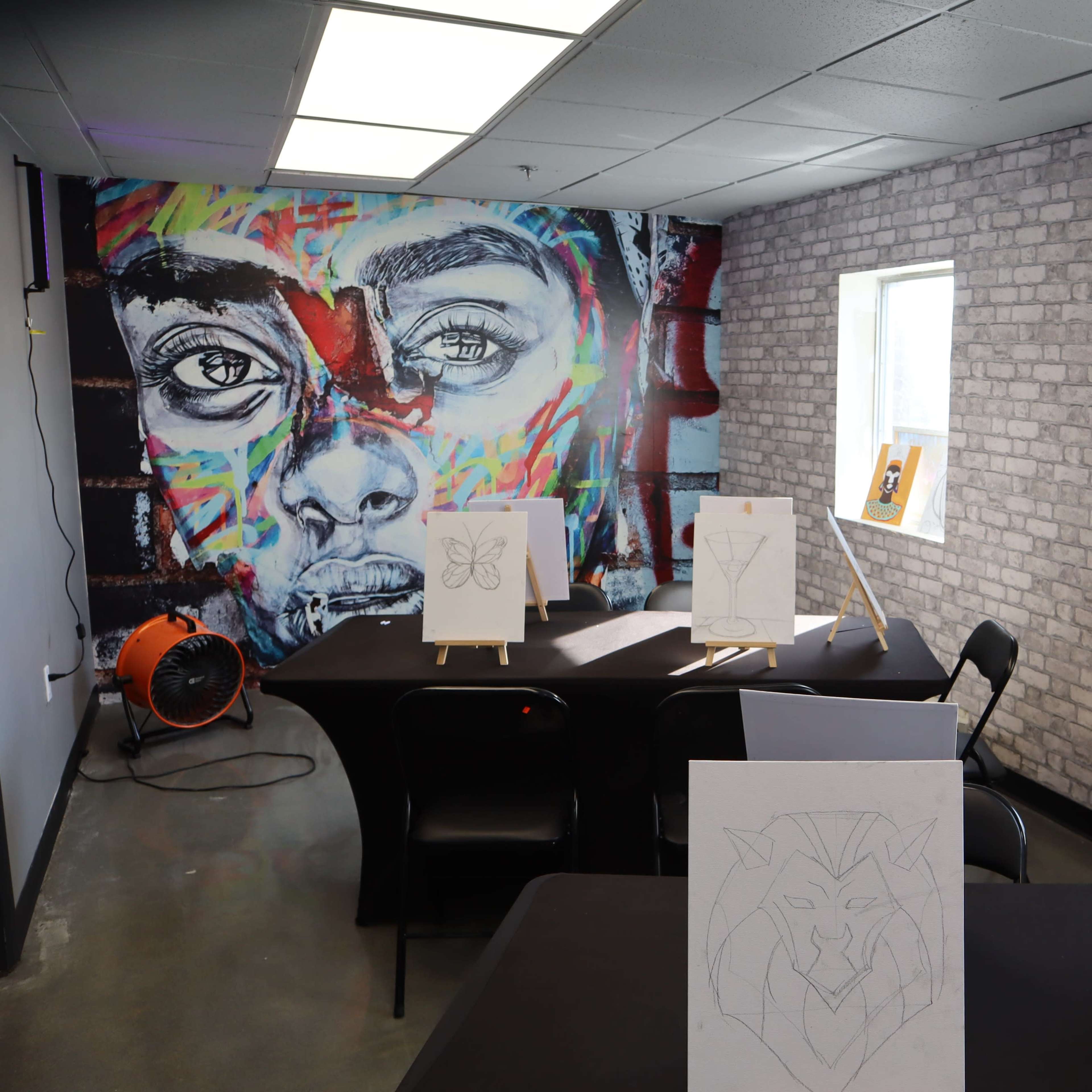 A spacious, brightly lit art room with a mural of a face on the brick wall and several easels displaying unfinished drawings.