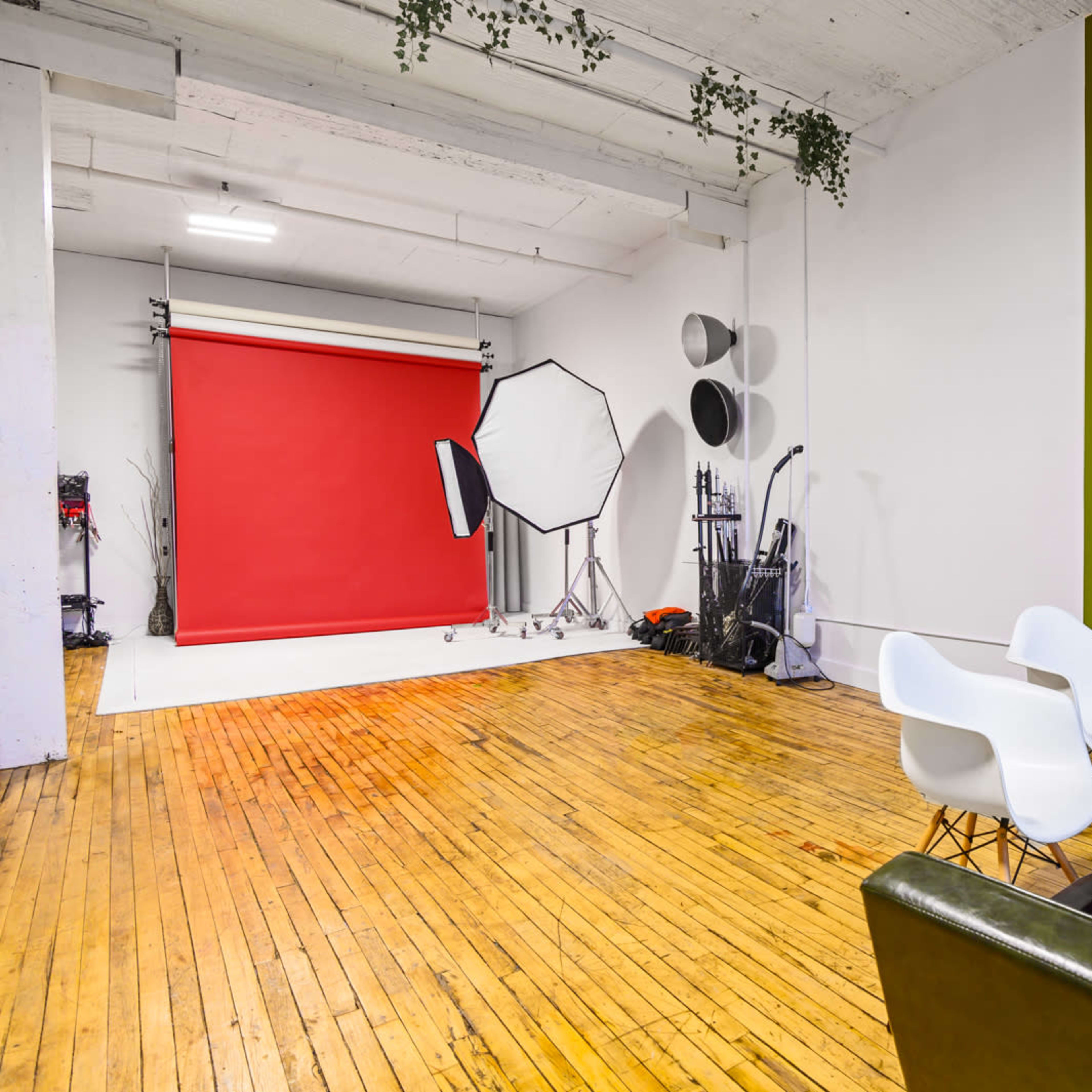 A photo studio with a red backdrop, softboxes, wooden floors, and a seating area featuring white chairs and a green wall.
