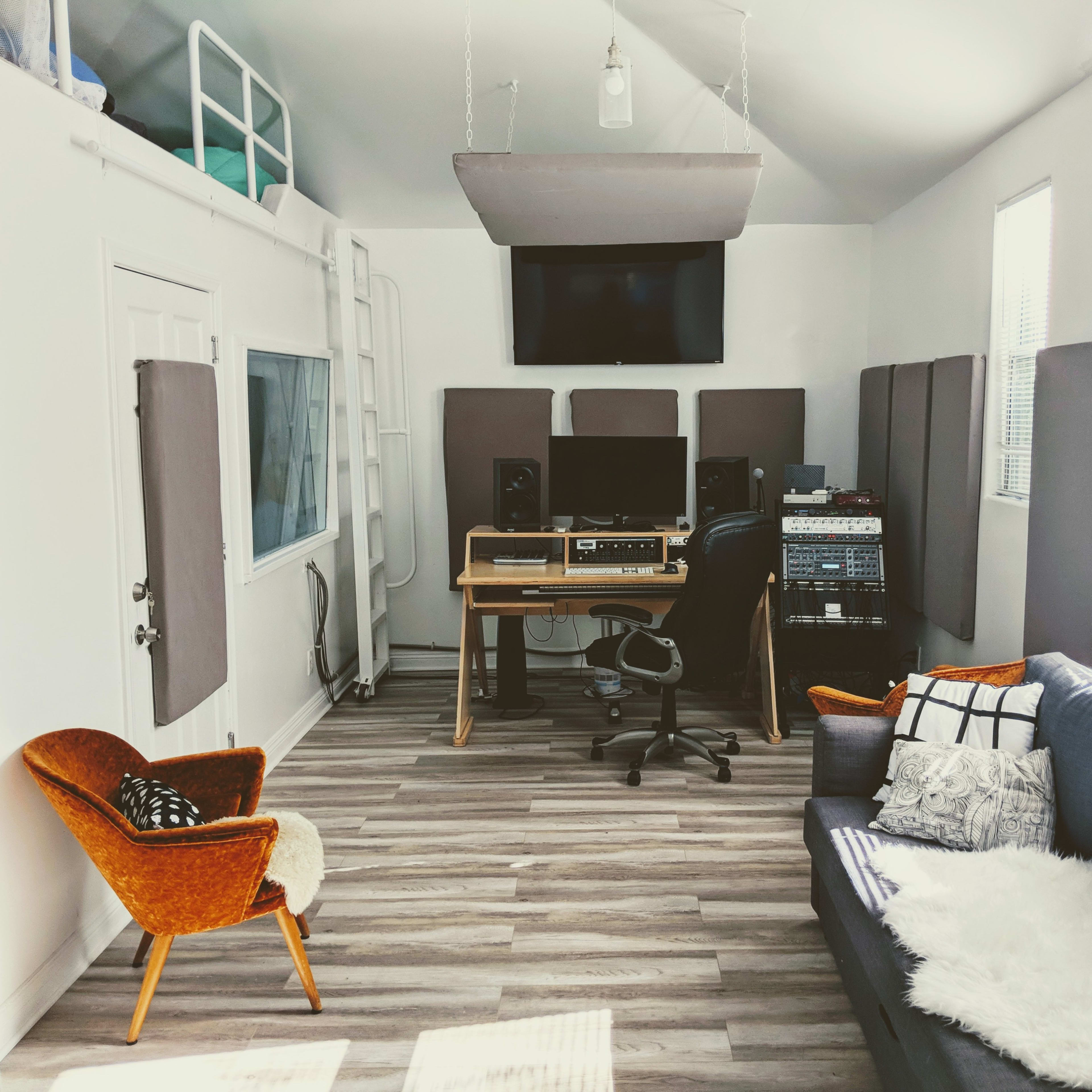 A cozy music studio featuring a desk with audio equipment, a comfortable couch, an armchair, and soundproof panels on the walls.
