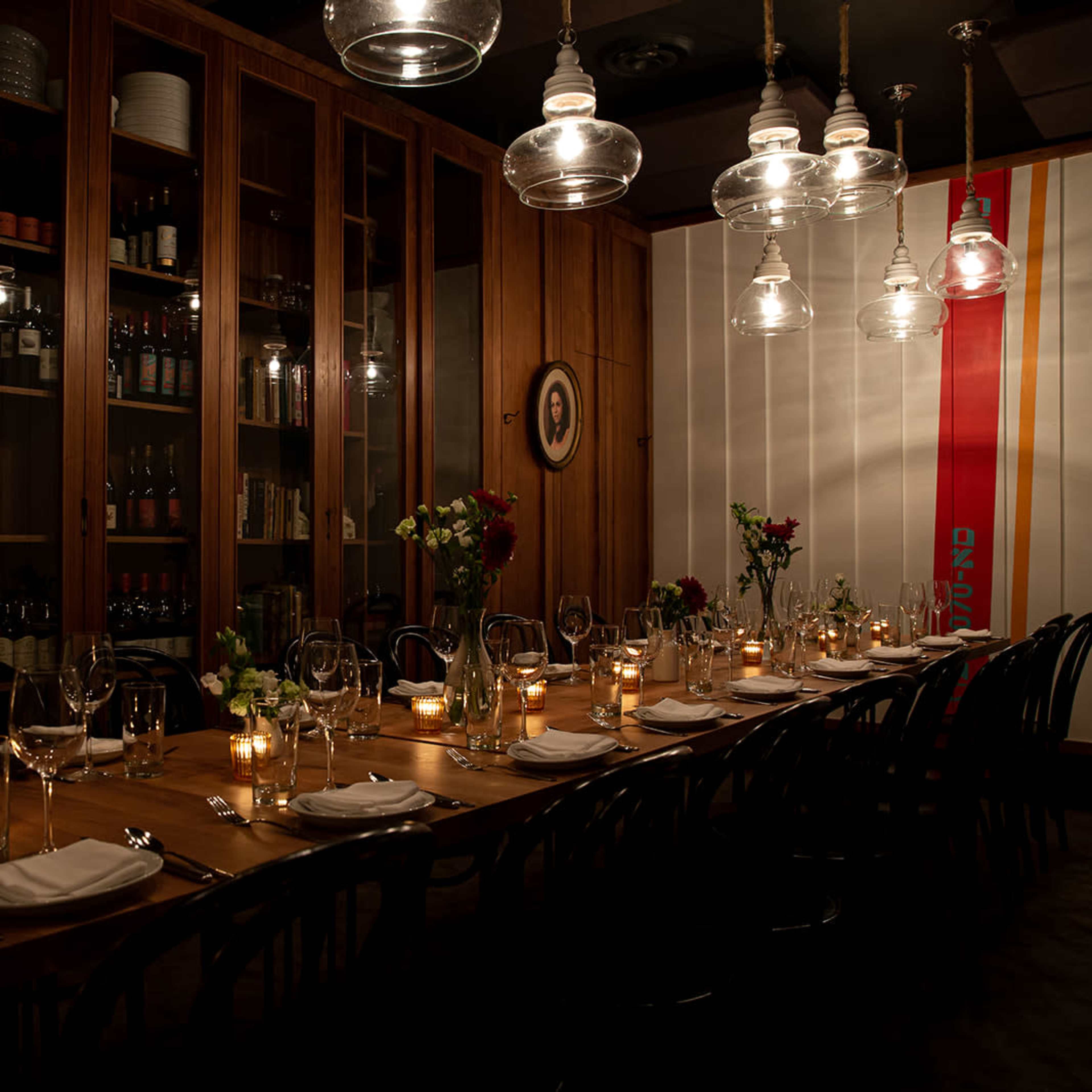 A long wooden dining table is set with candles and flower arrangements, surrounded by shelves stocked with bottles in a warmly lit restaurant.