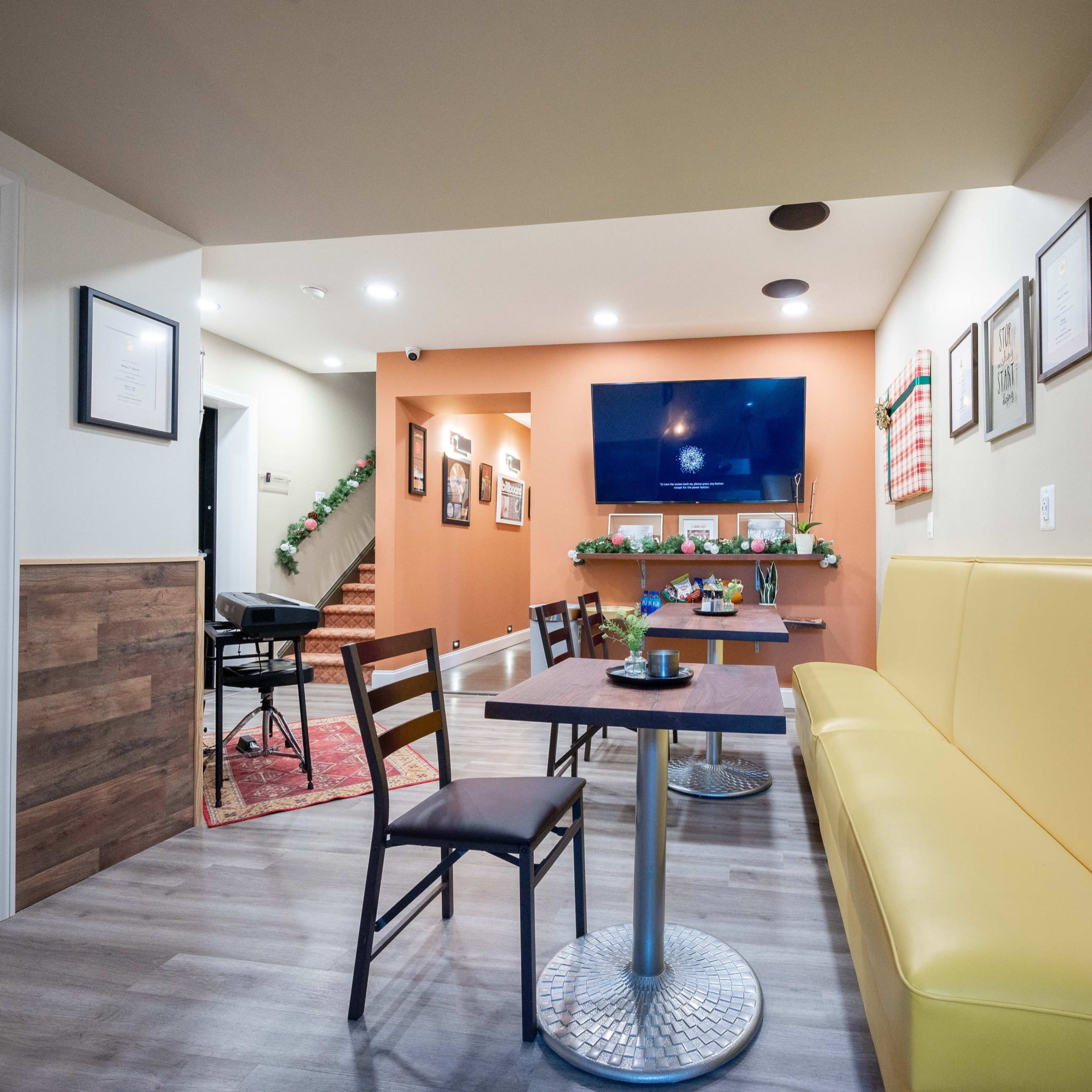 A cozy café interior featuring a yellow banquette, a small table with a chair, and a wall-mounted television, with stairs visible leading to another area.