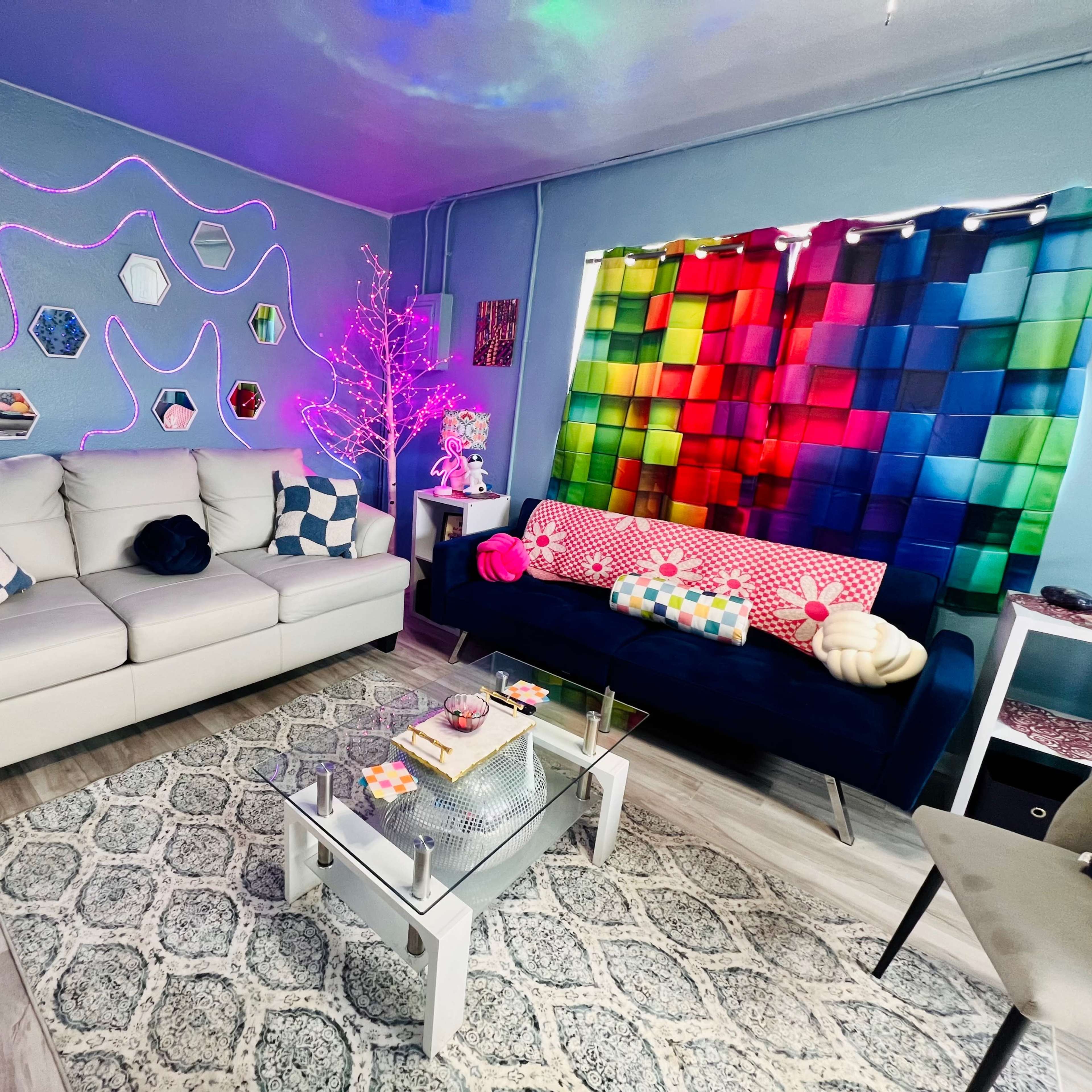 A colorful living room with a white sofa, a dark sofa adorned with cushions, a glass coffee table, and decorative wall art illuminated by colorful lights.