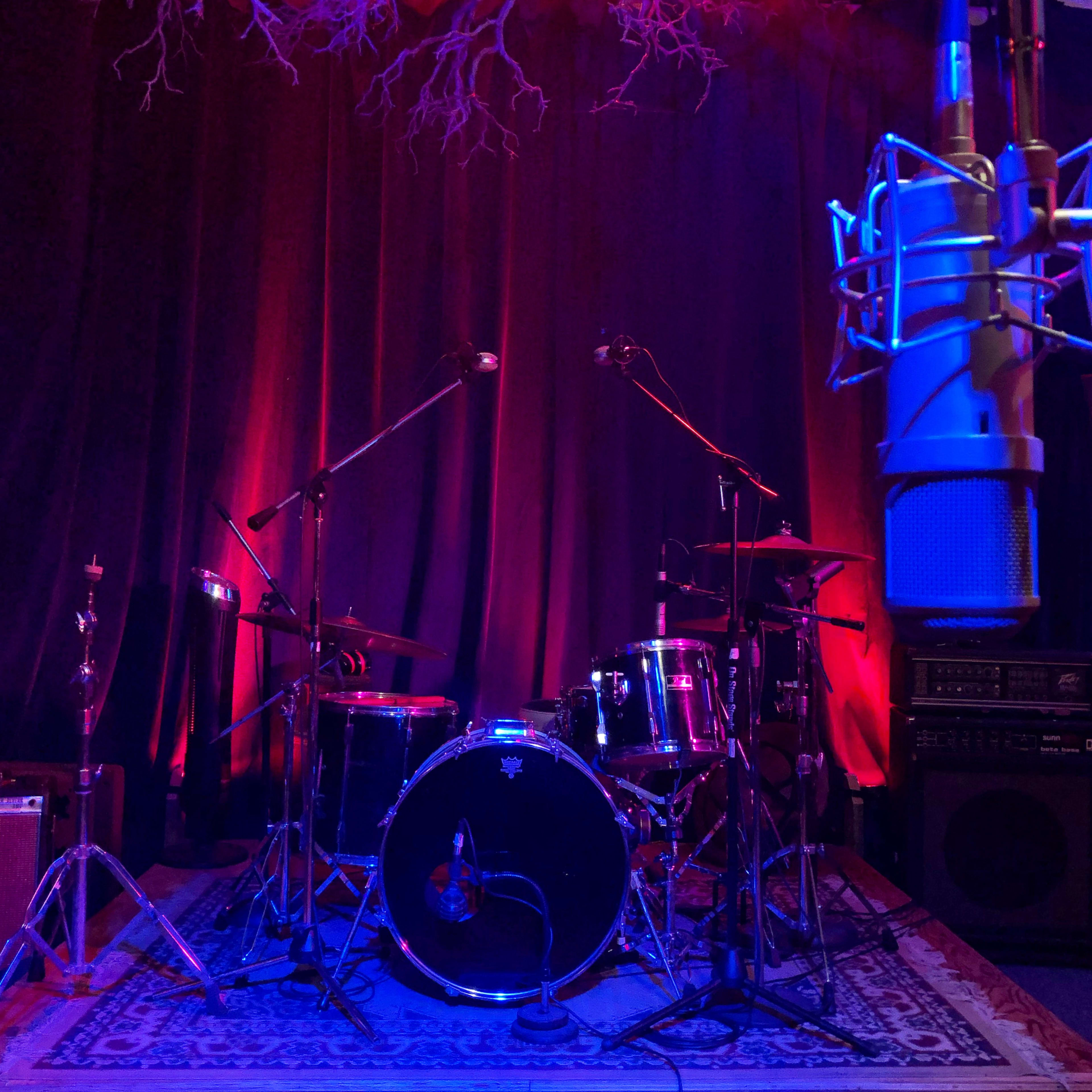 A drum set is positioned on a carpet in a studio space with a large microphone and curtain backdrop illuminated by colored lights.