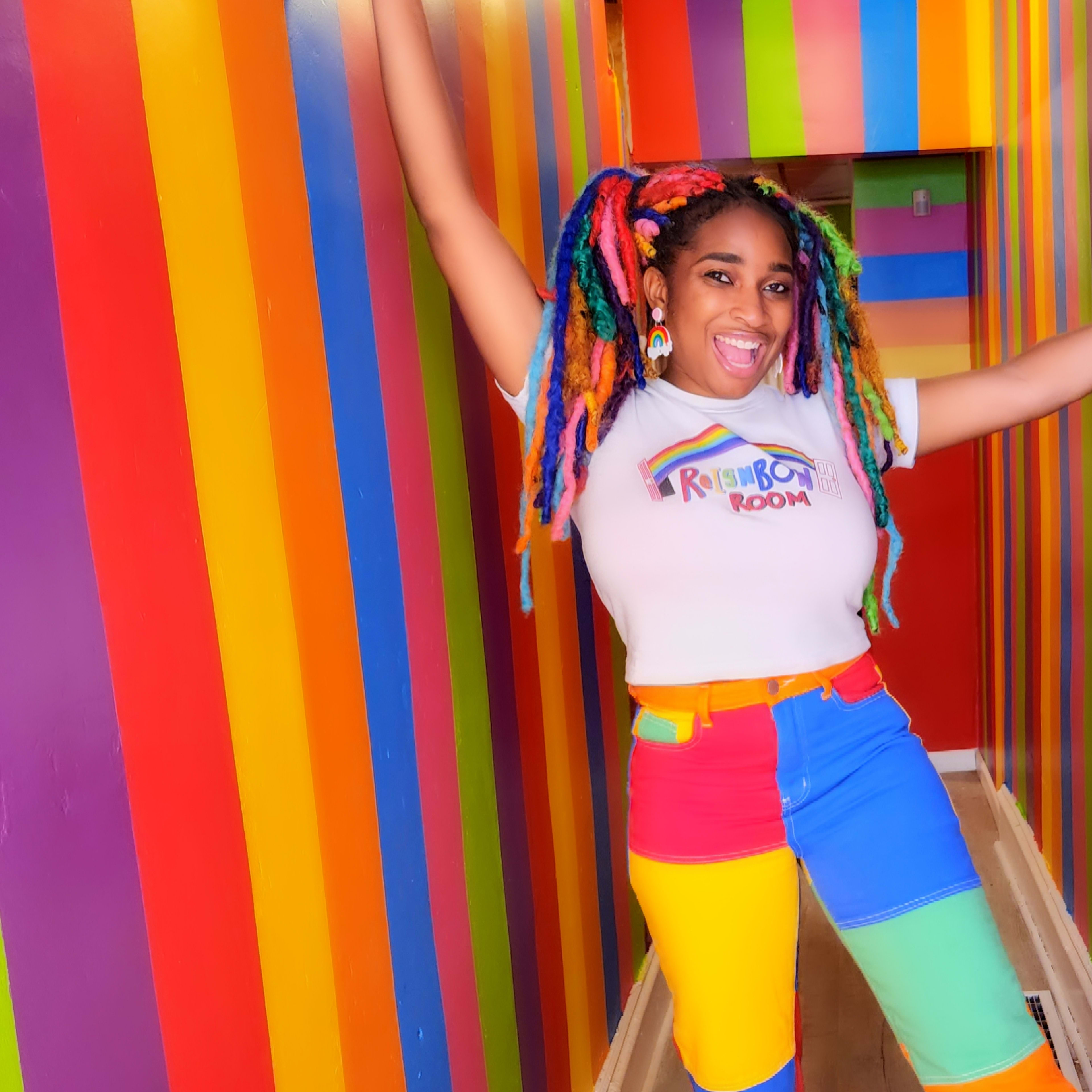 A person with colorful, multi-colored hair poses in a brightly painted, striped hall.