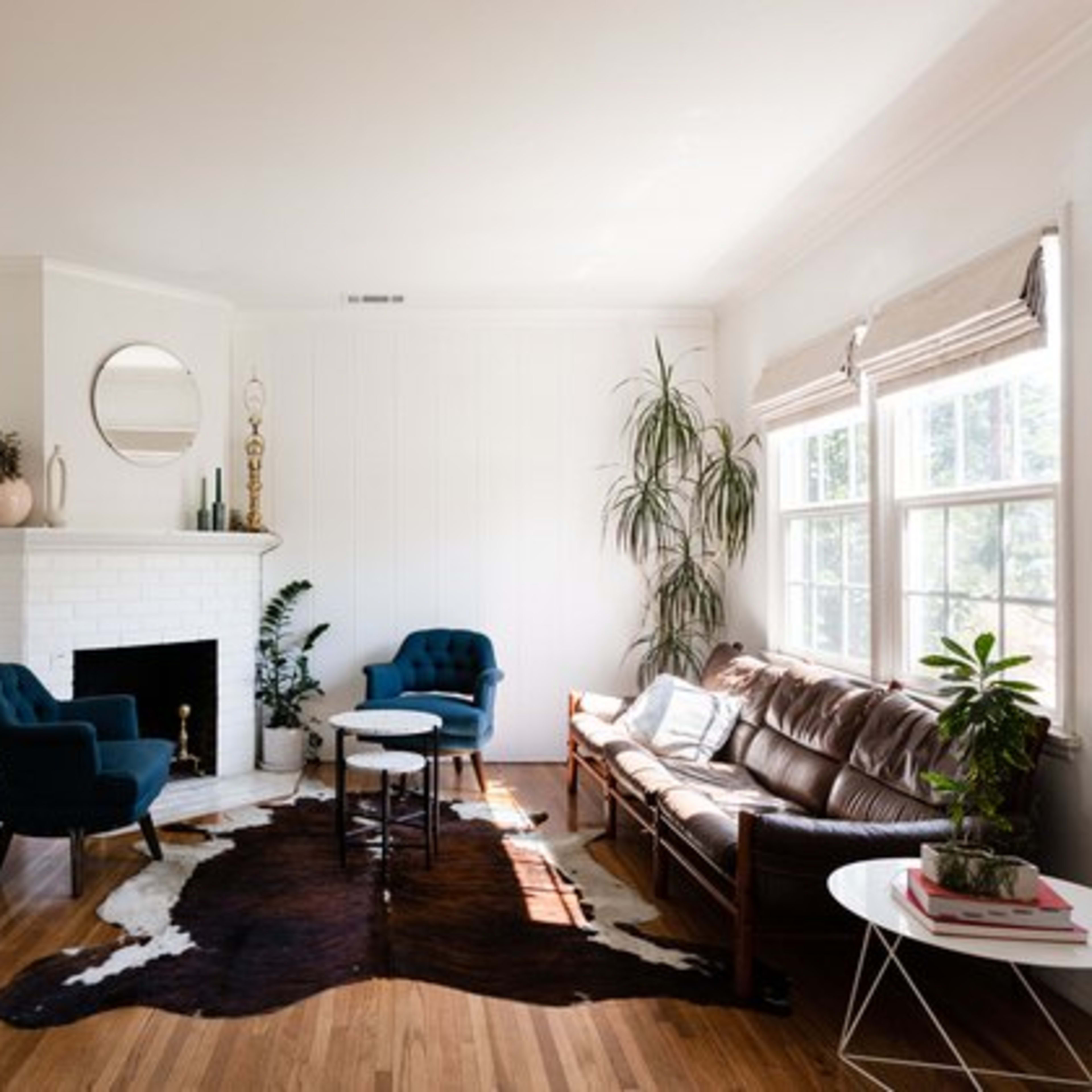 A stylish living room features a brown leather sofa, two blue chairs, a round coffee table, and a white fireplace with a circular mirror above it.