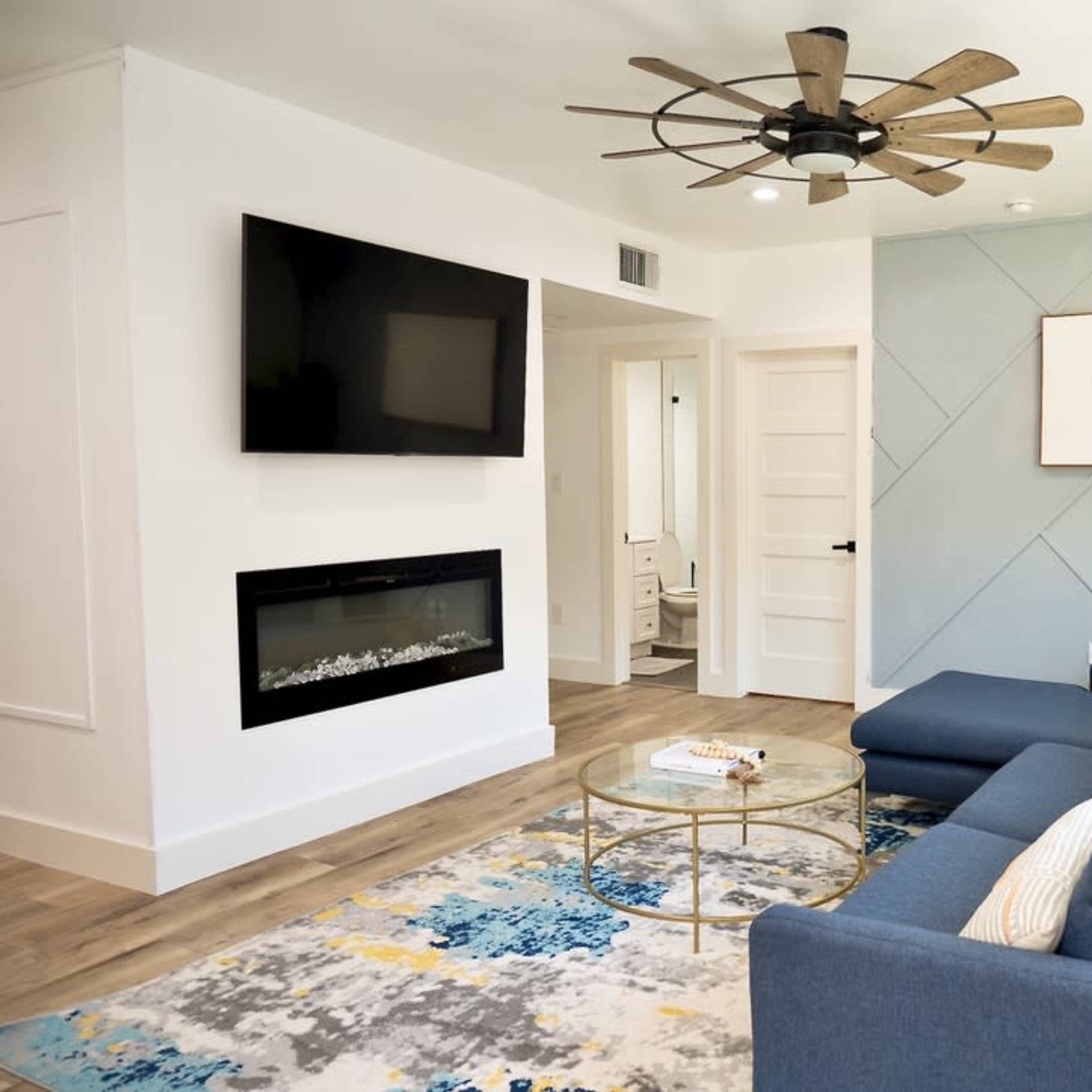 A modern living room with a blue sectional sofa, a wall-mounted television, an electric fireplace, and a coffee table on a patterned rug.