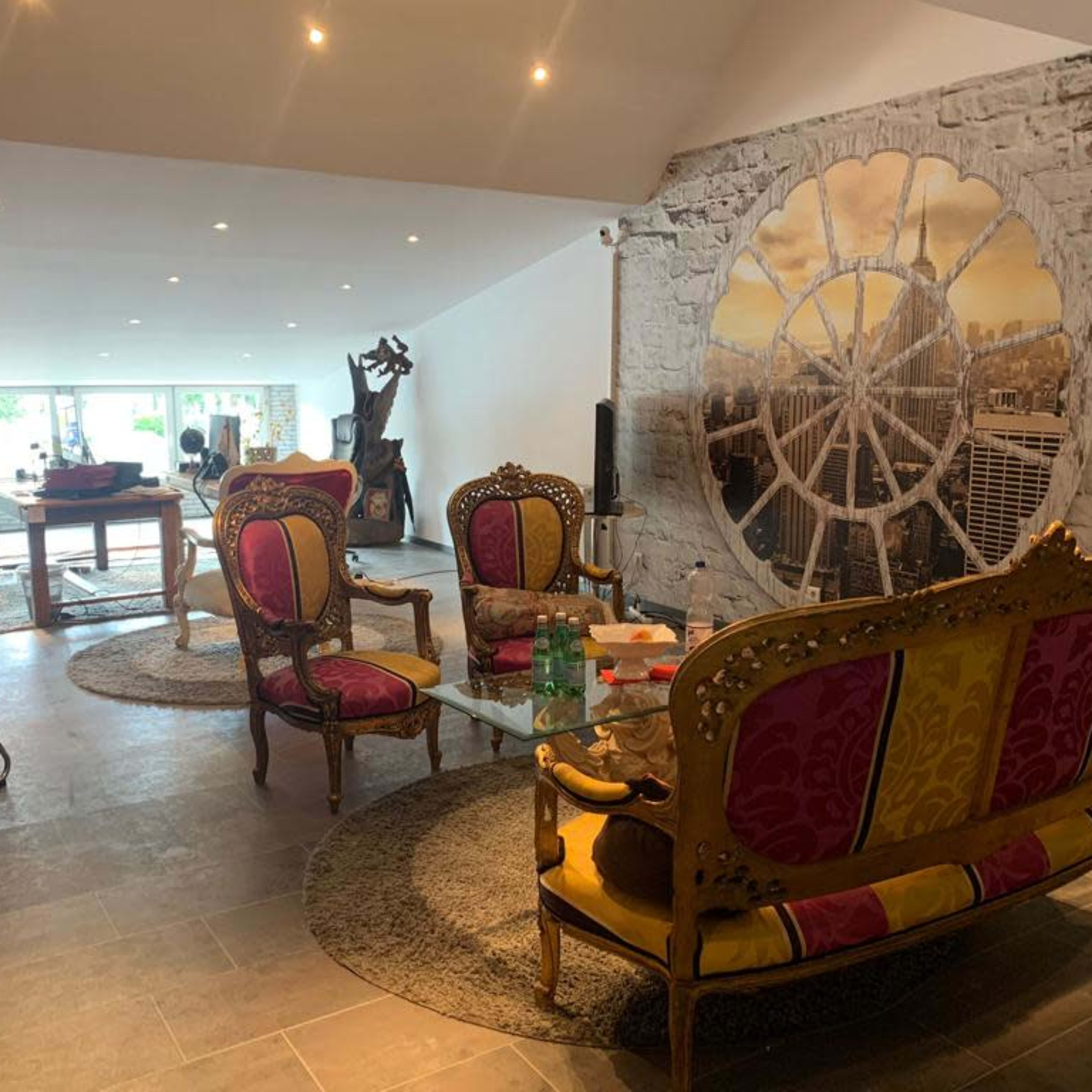 The room features elaborate, colorful chairs arranged around a glass table, with a large decorative mural and a stone wall as the backdrop.