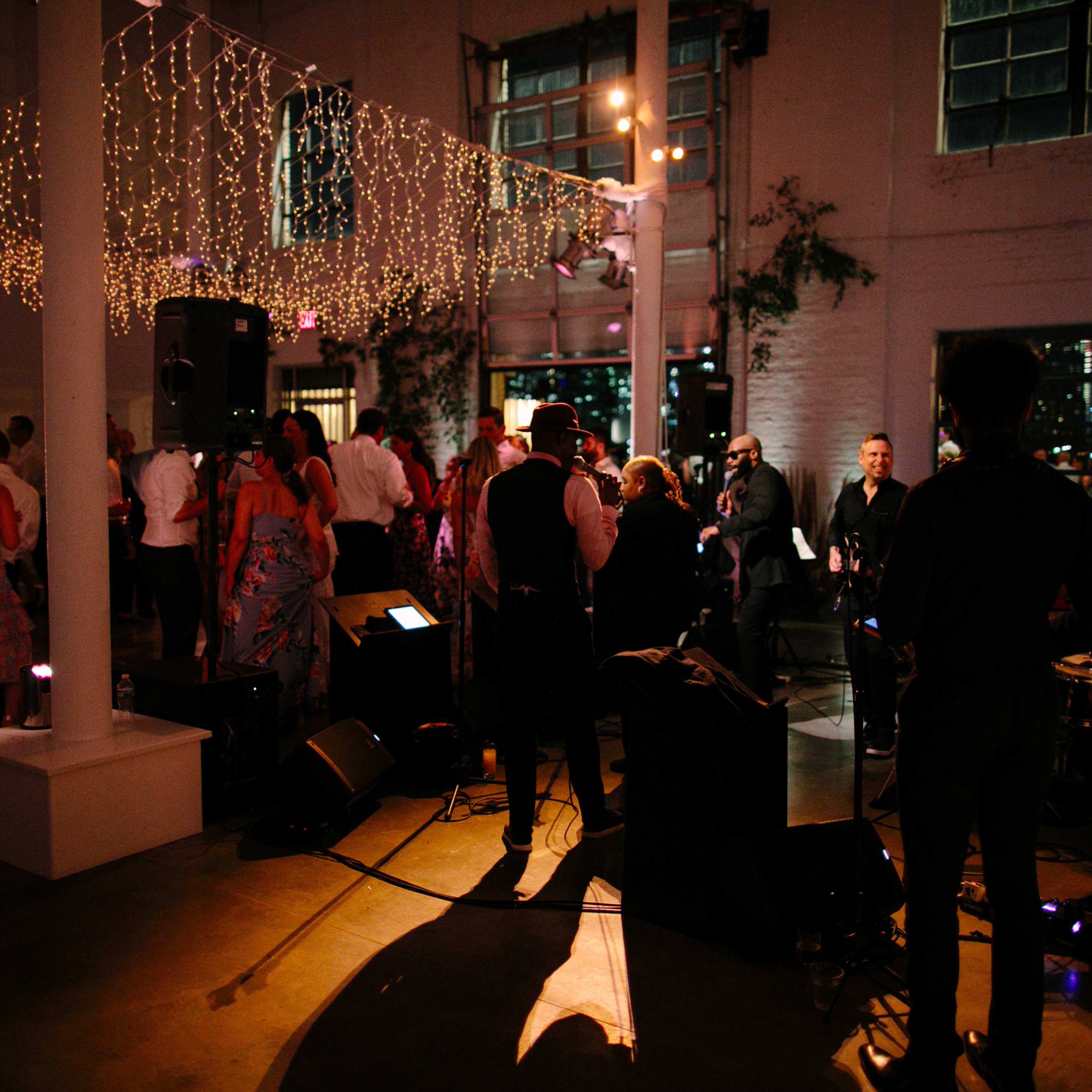 A lively event takes place in a spacious venue adorned with string lights, featuring a band performing while guests dance and socialize.