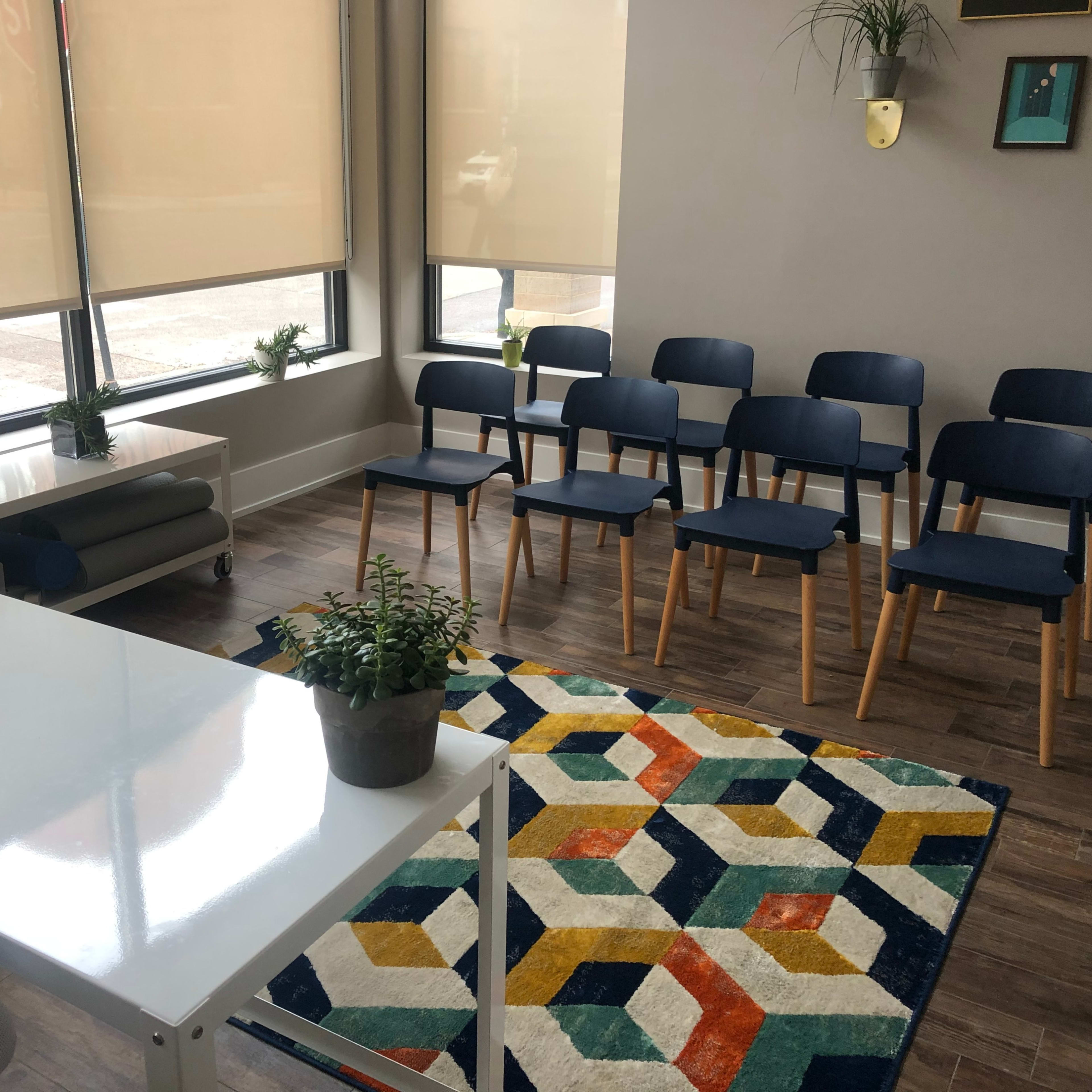 A modern waiting area with a white desk, several blue chairs arranged in rows, and a colorful geometric rug.