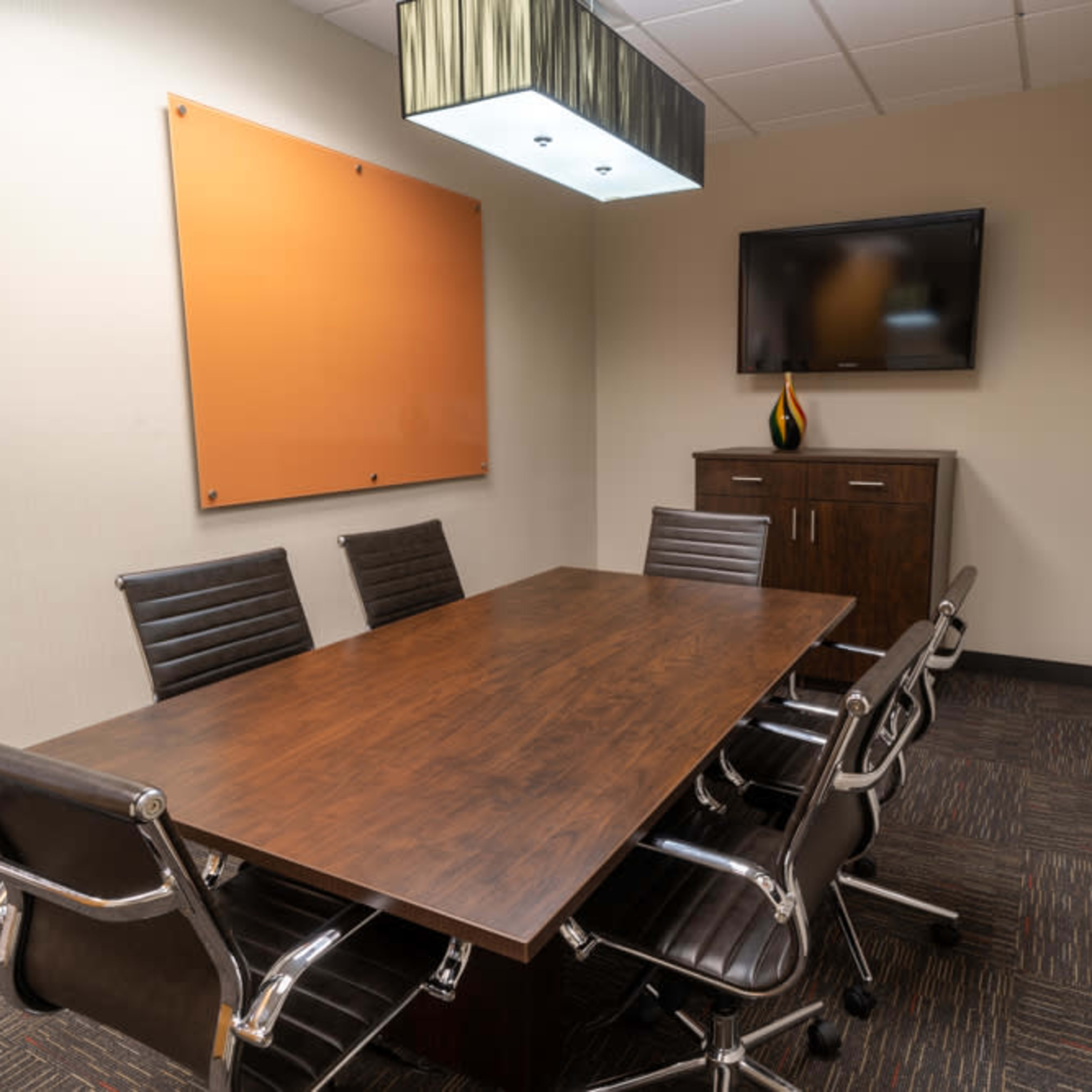A conference room features a long wooden table surrounded by black leather chairs, a mounted television, and wall art.