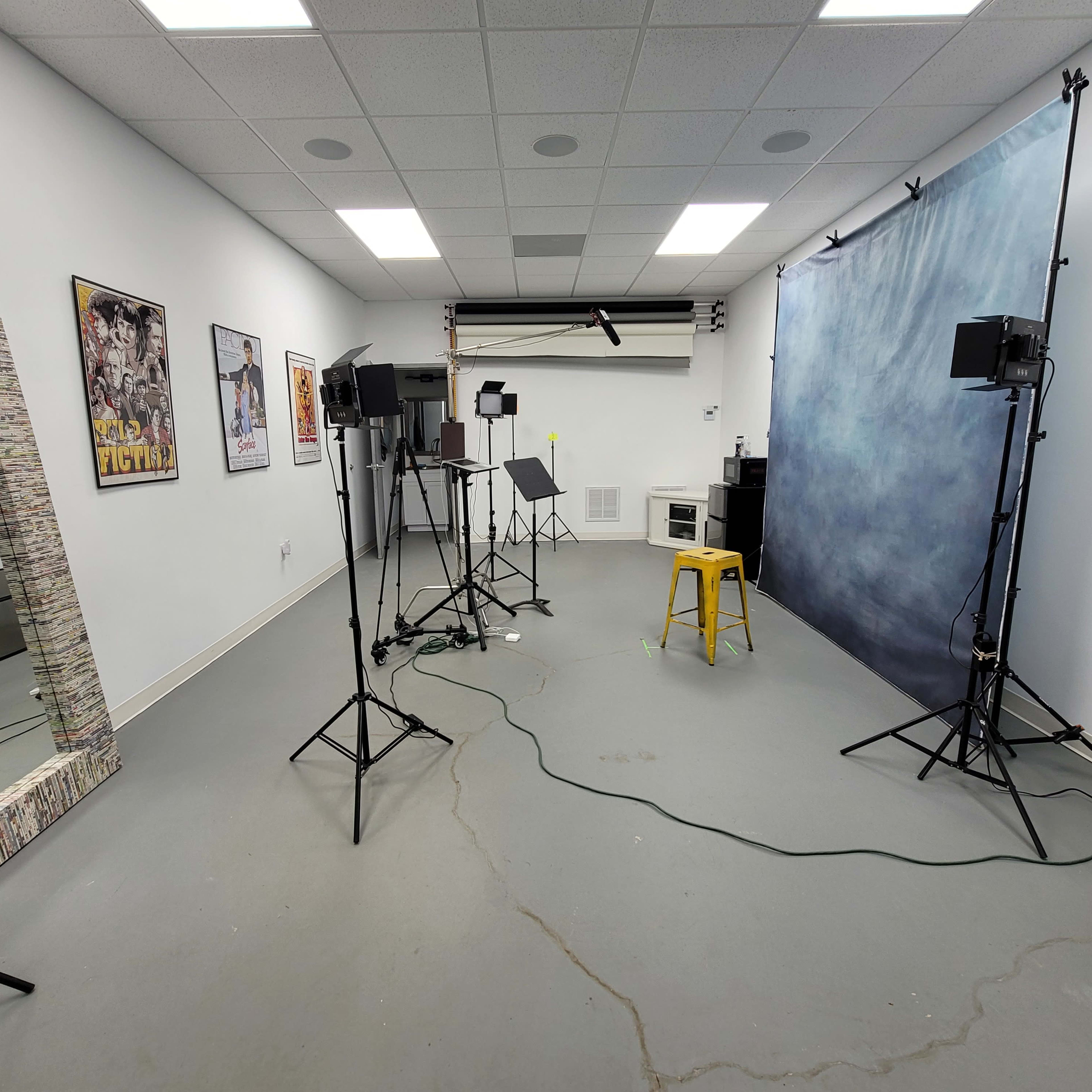 A photography studio with a blue backdrop, lighting equipment, a yellow stool, and a mirror on the wall.