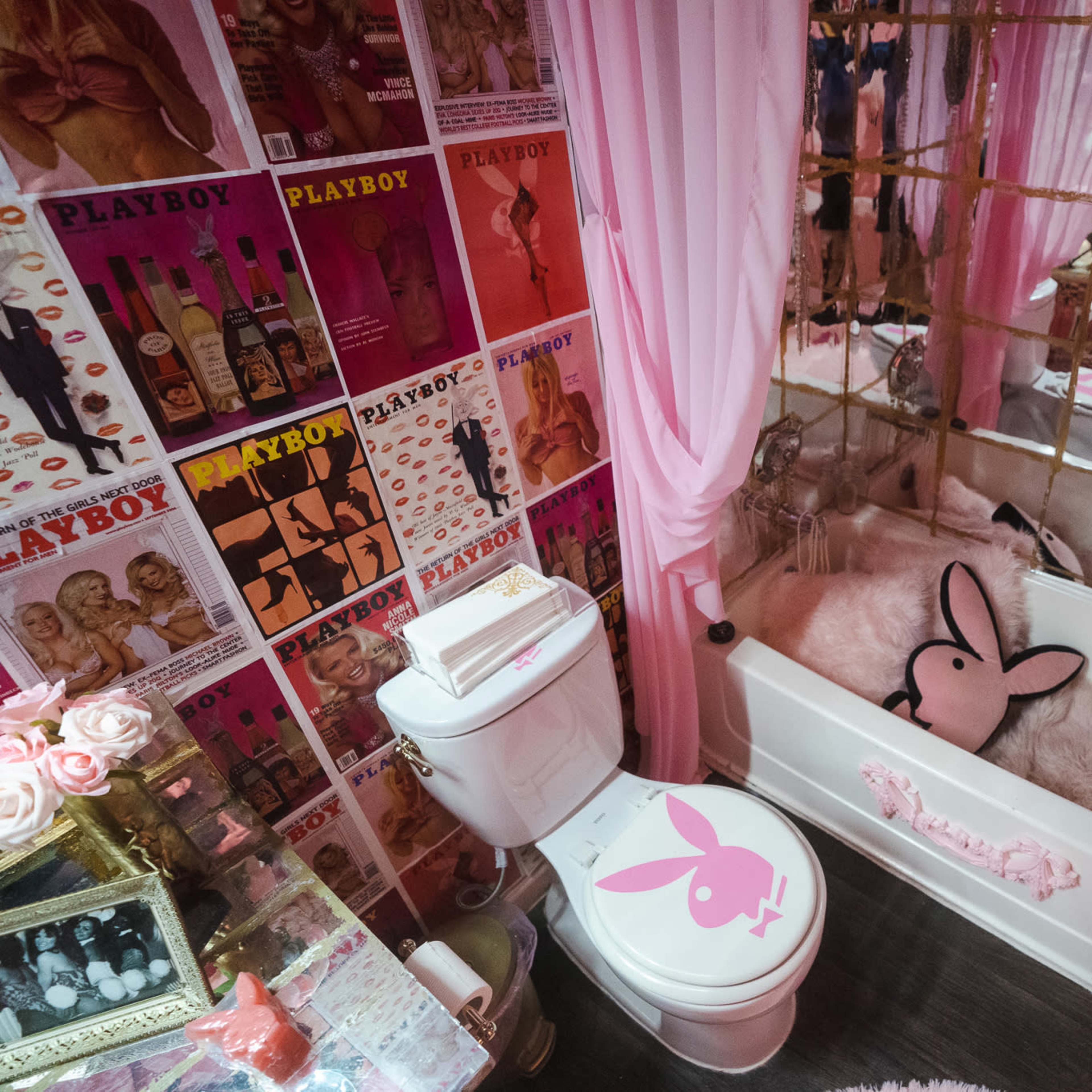 A bathroom decorated in pink tones with Playboy-themed posters on the walls, a plush bunny-shaped pillow in the bathtub, and a matching toilet seat cover.
