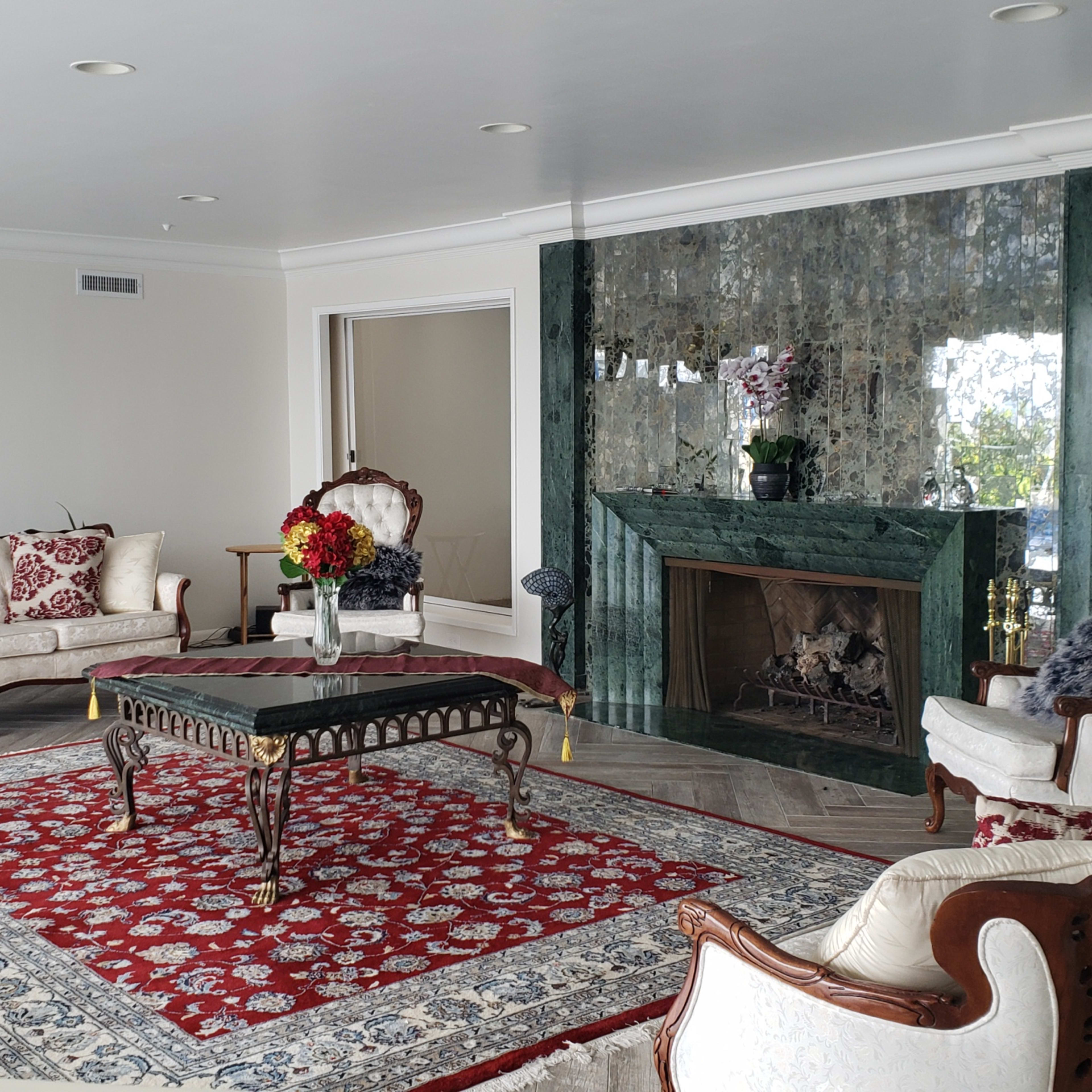 A spacious living room with cream-colored sofas, a red patterned rug, and a green marble fireplace with a mirrored wall.