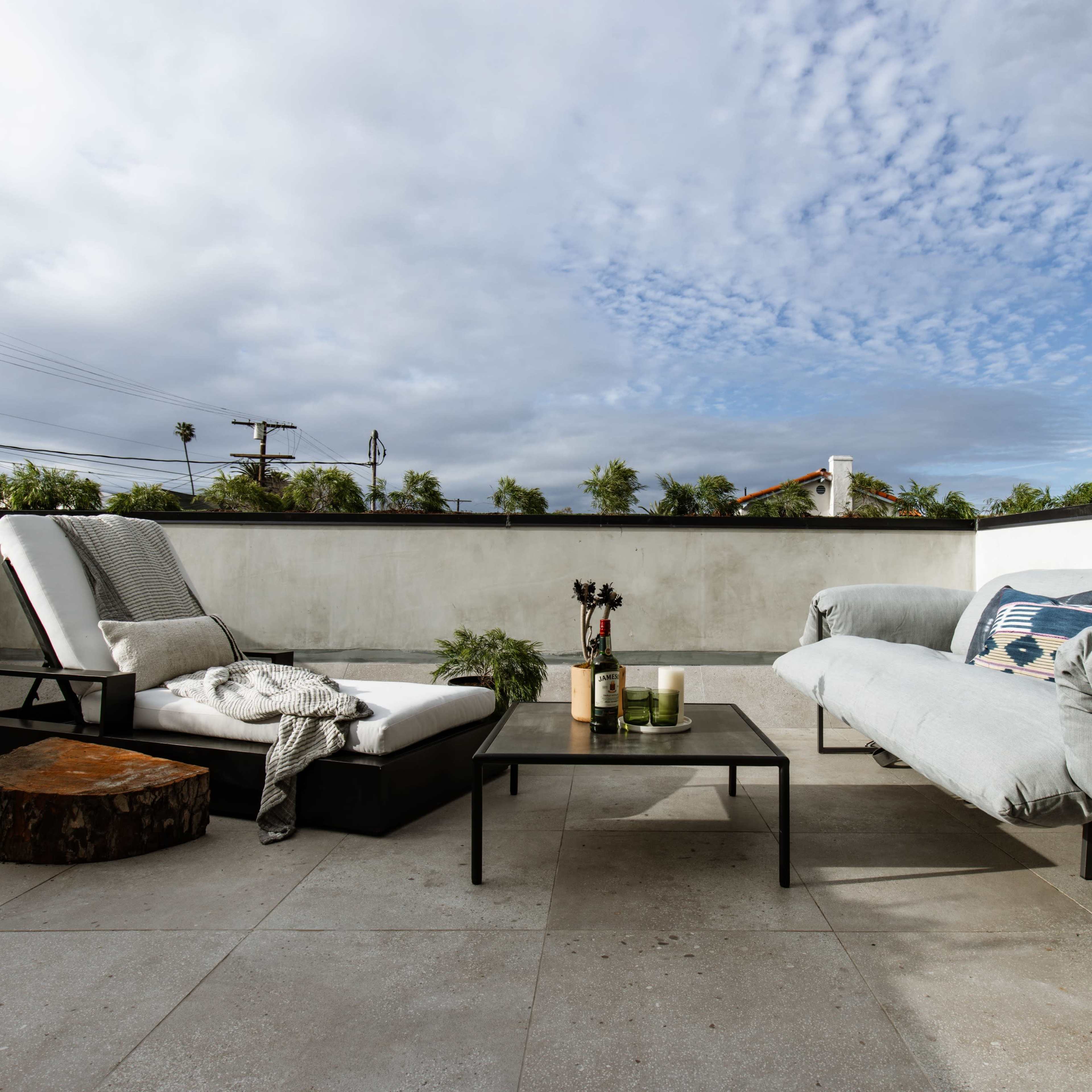 A rooftop terrace with two lounge chairs, a coffee table, and a wooden stump amidst a cloudy sky and greenery in the background.