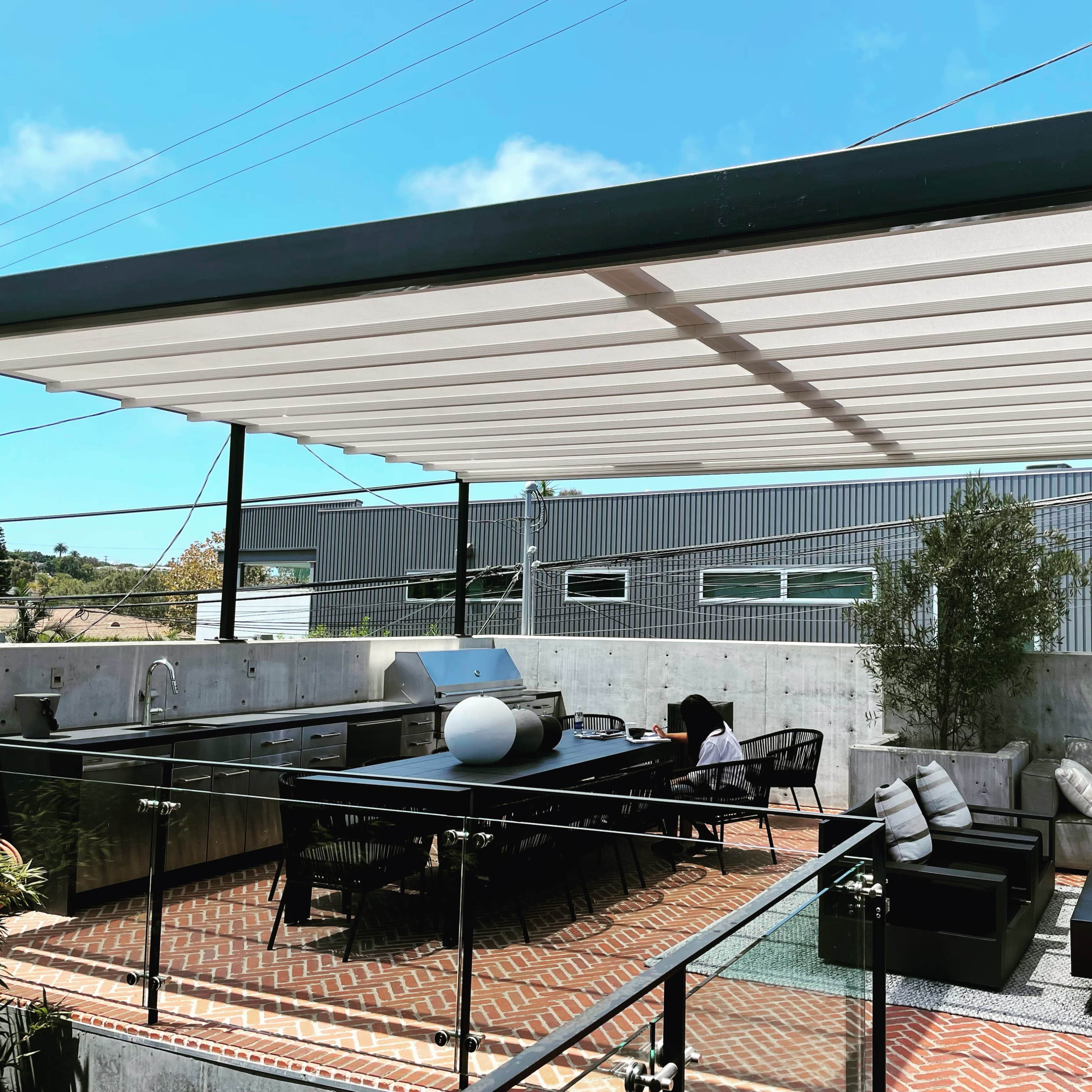 A modern rooftop terrace featuring a dining area with a large table, comfortable seating arrangements, and a grill under a slatted canopy.