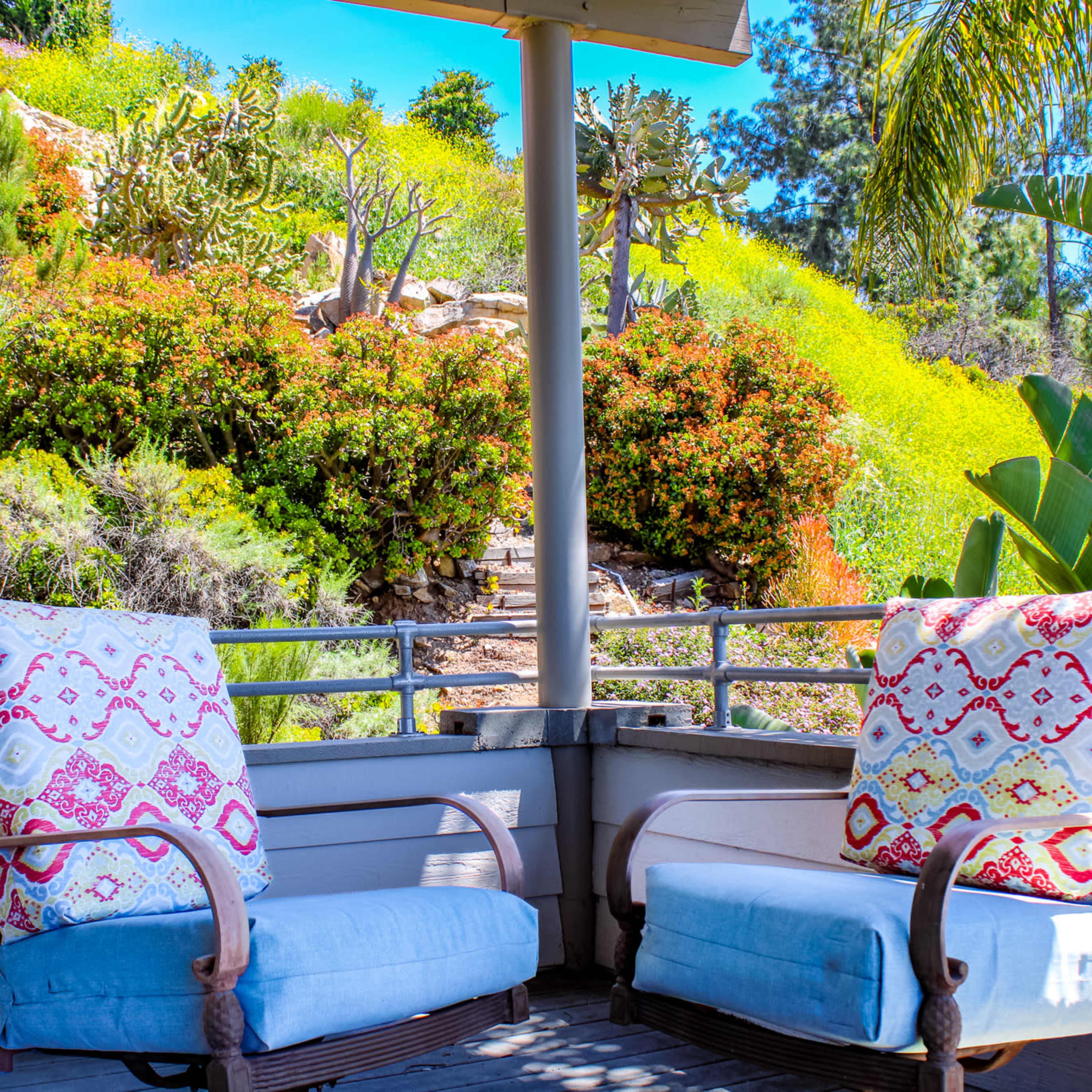 Two cushioned chairs with decorative pillows sit on a wooden patio, overlooking a lush hillside with greenery and colorful plants.