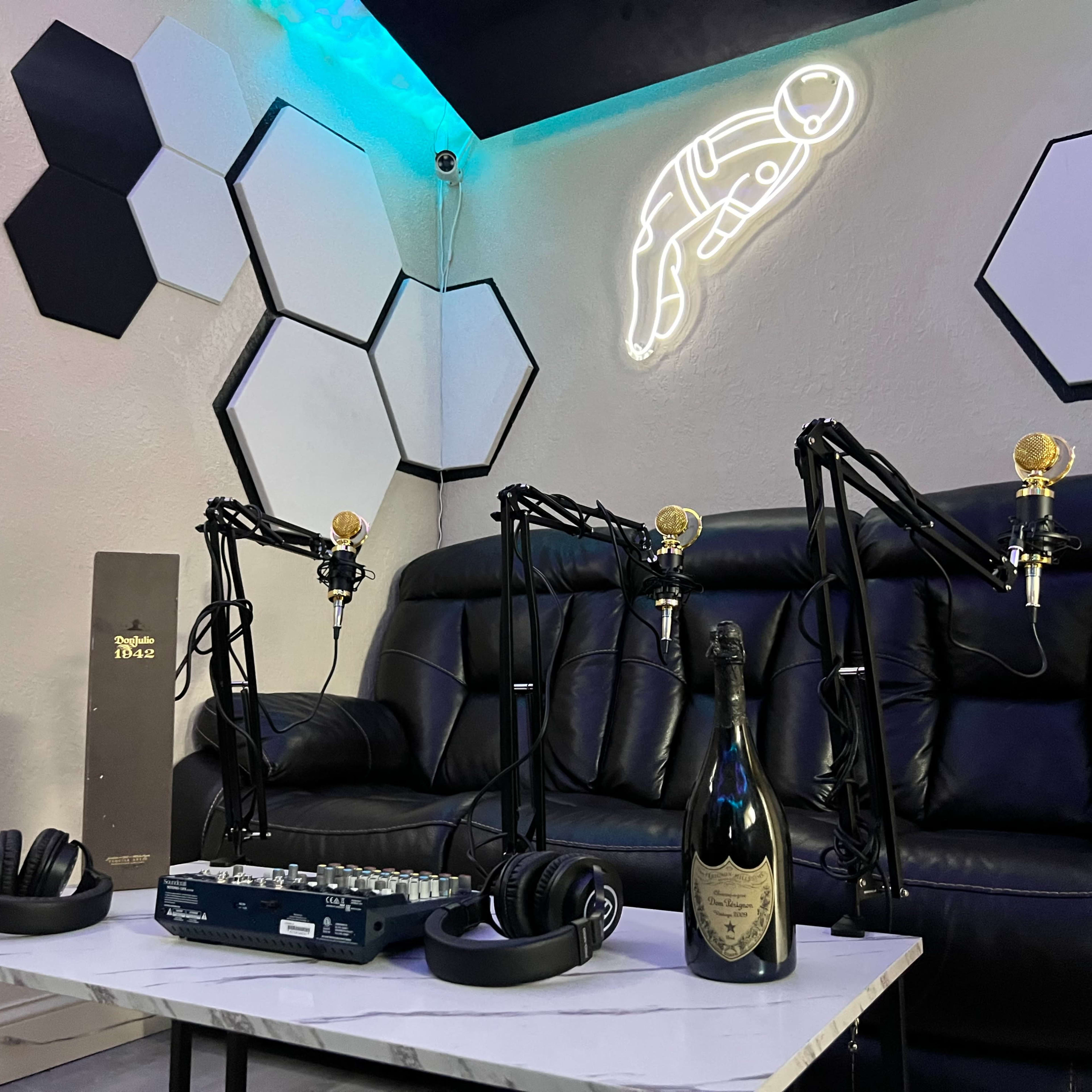 A podcast studio setup with three microphones on stands, a black couch, headphones, a sound mixer, and a bottle of champagne on a white table, all against a backdrop of hexagonal wall decorations and neon lights.
