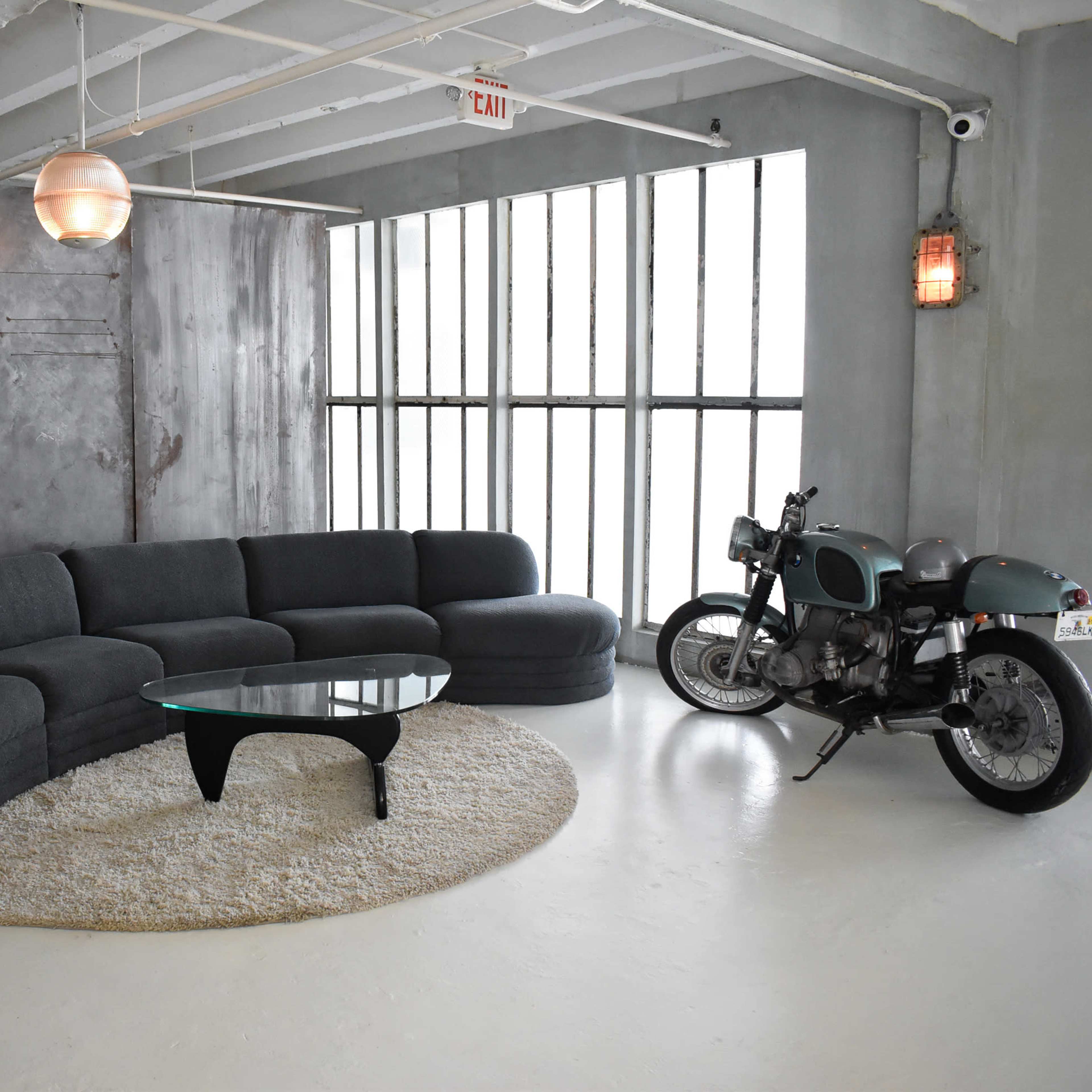 A modern living room features a curved gray sofa, a round glass coffee table on a circular rug, and a vintage motorcycle parked beside a large window.