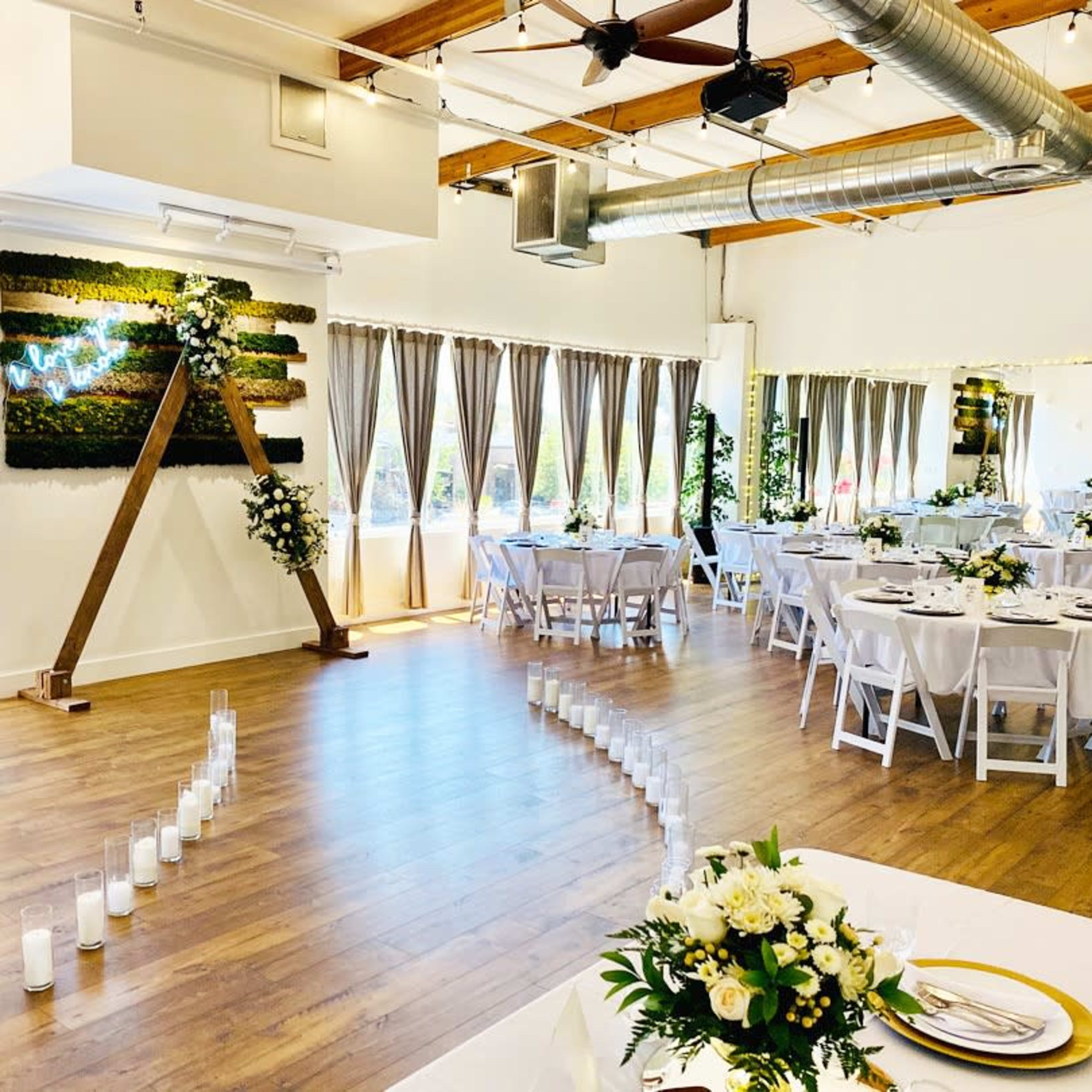 An elegantly decorated event space with tables set for a gathering, featuring a floral arrangement and a walkway lined with candles.