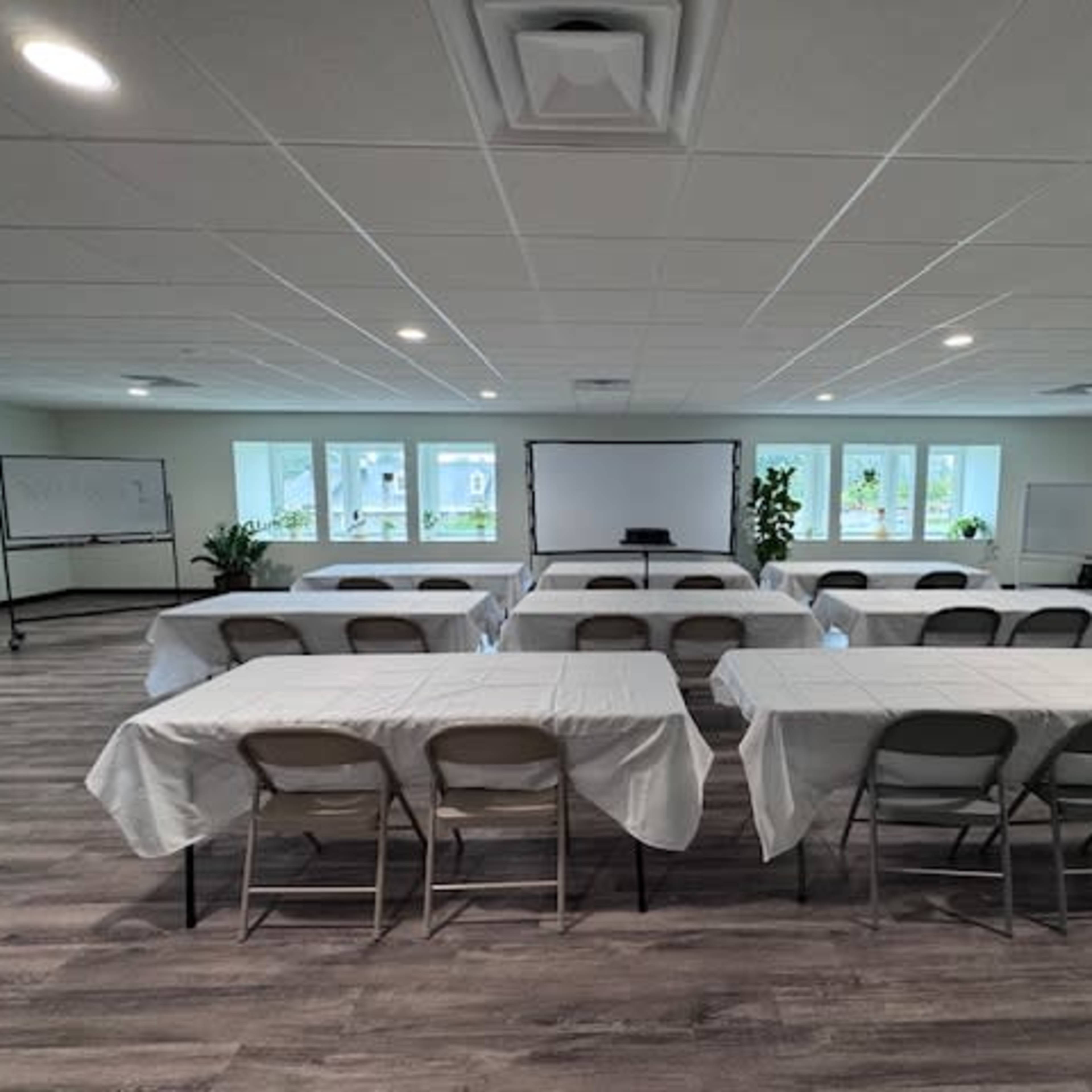 A spacious room set up for a meeting, with rows of tables covered in white tablecloths and chairs arranged in front of a projector screen.