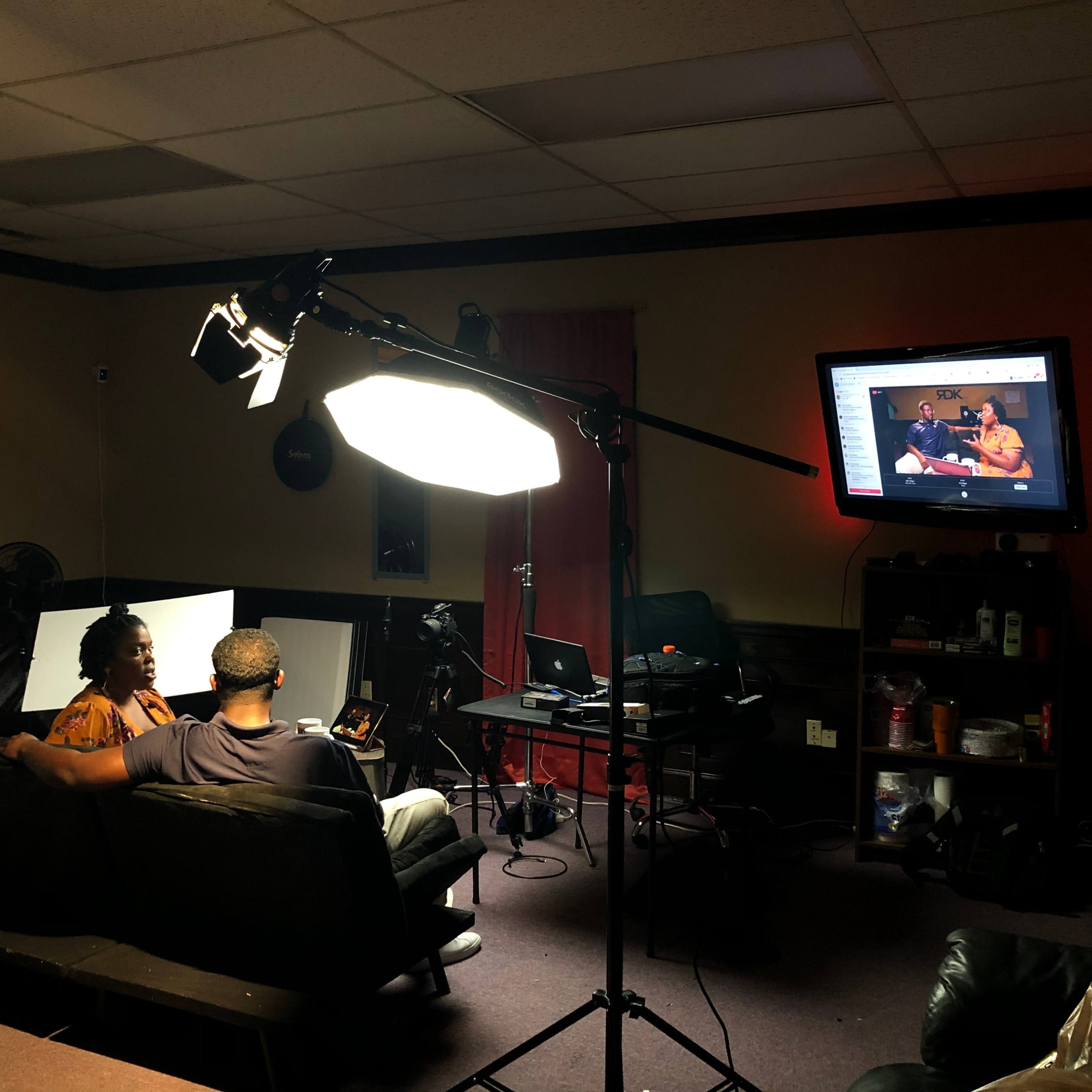 A person sits on a couch in a dimly lit room, facing a television screen while another person is seated nearby, surrounded by filming equipment.