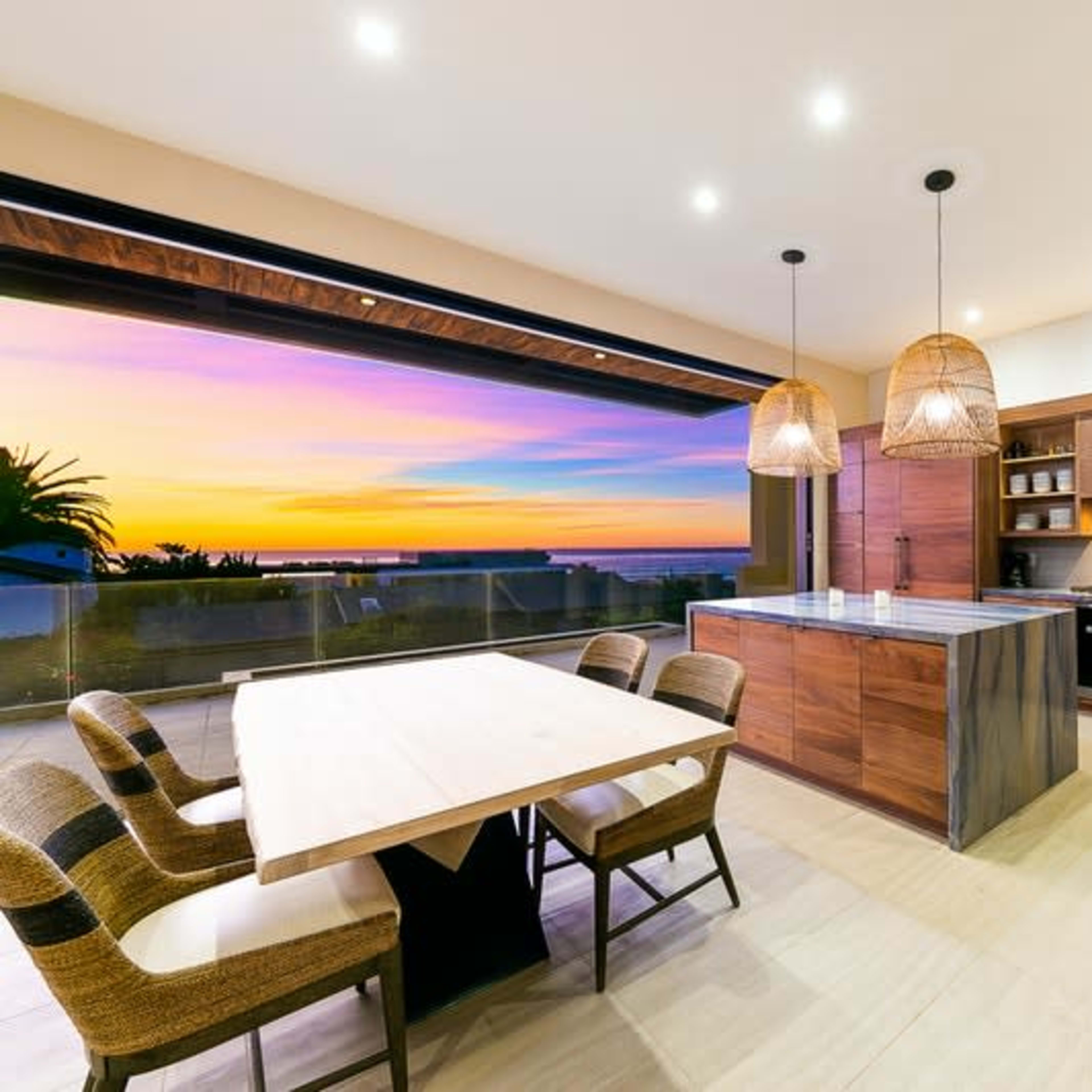 A modern kitchen features a large island, wooden cabinets, and a dining table with a view of a colorful sunset through expansive windows.