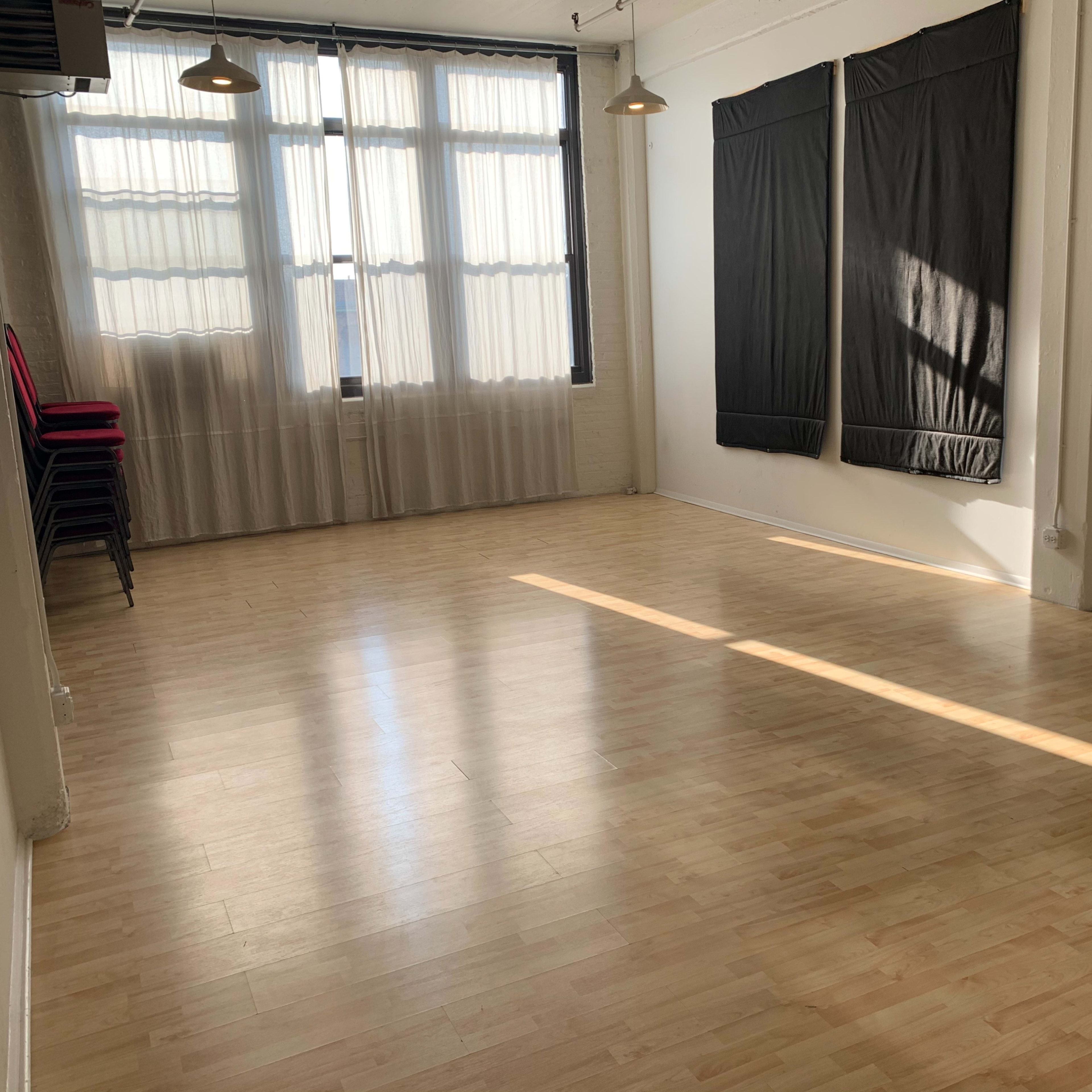 A spacious, sunlit room with wooden flooring and black fabric-covered walls, featuring stacked chairs in one corner.