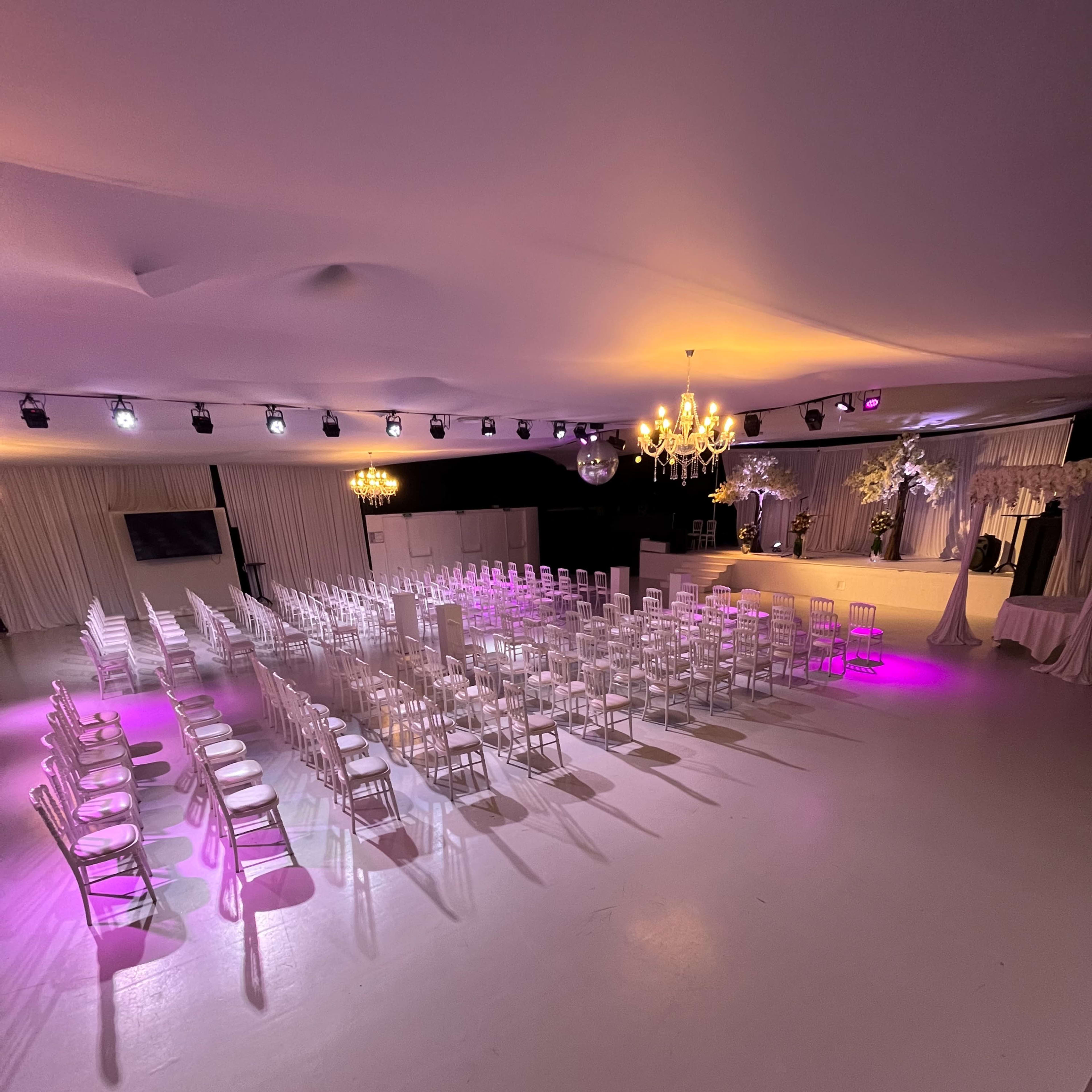 A spacious event venue features rows of white chairs set up for a gathering, illuminated by pink lights and a chandelier overhead.