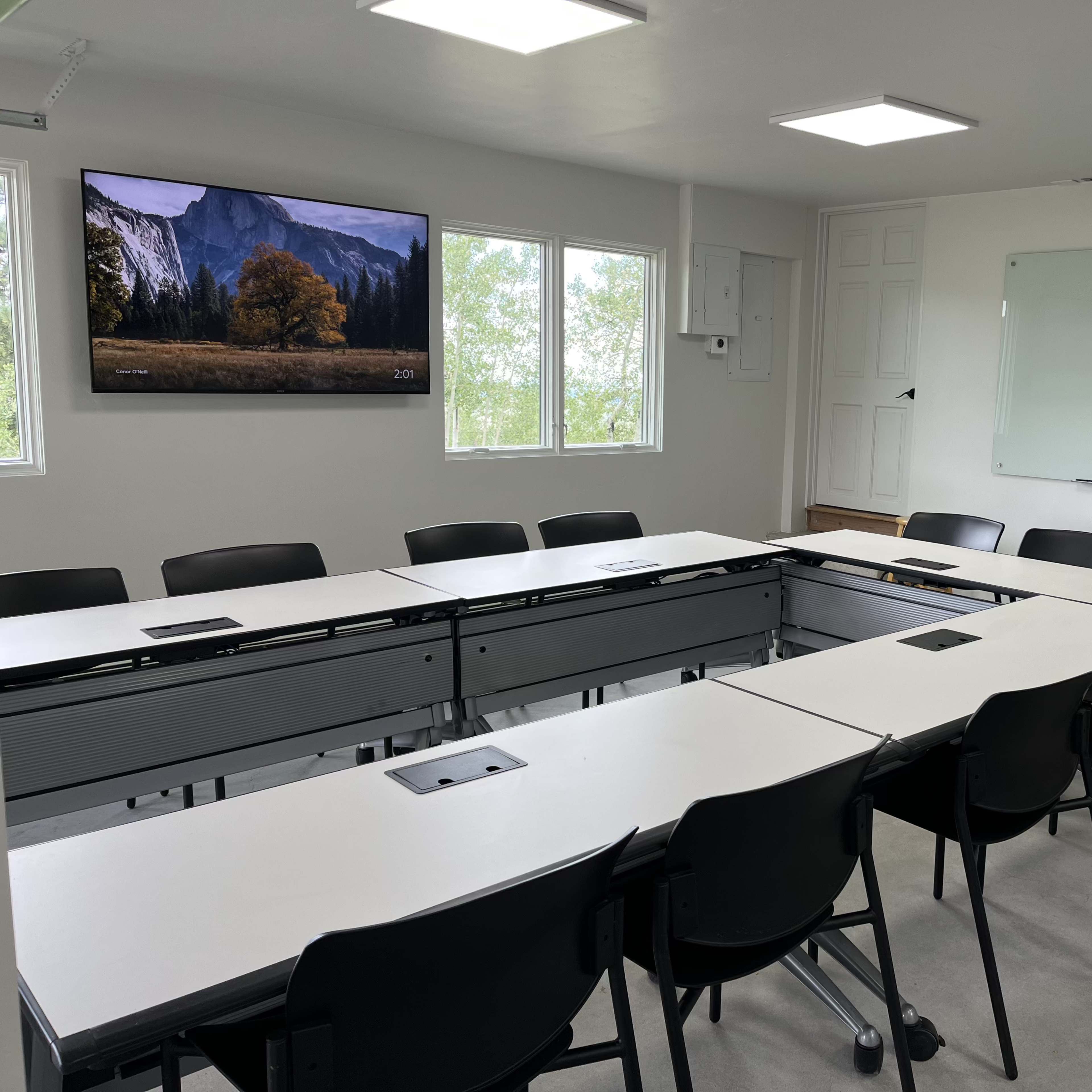 A modern conference room with a large screen, multiple whiteboards, and a setup of tables arranged in an oval shape surrounded by black chairs.