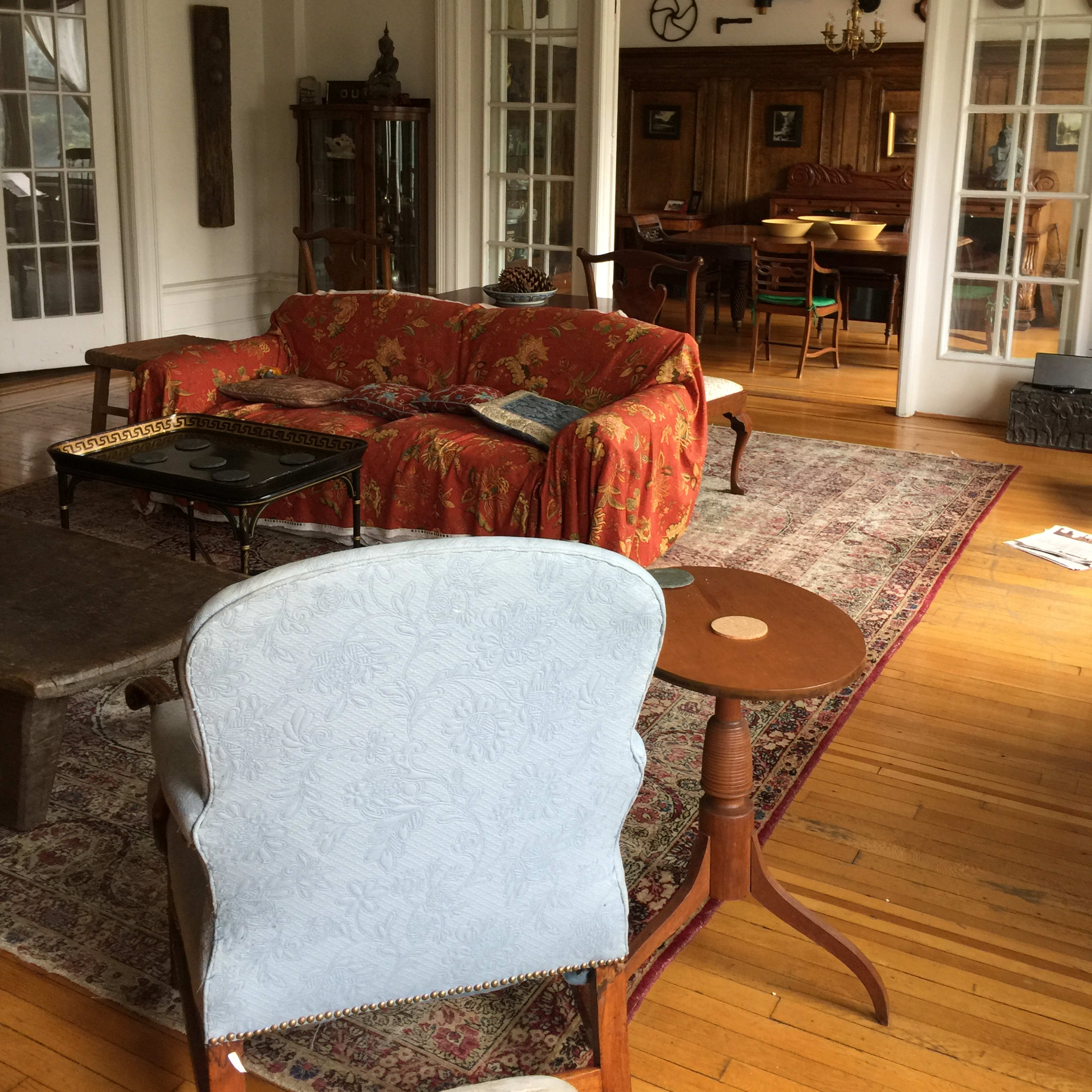 A spacious, elegantly furnished living room featuring a red floral sofa, a wooden coffee table, and a dining area in the background with several chairs and a wooden sideboard.