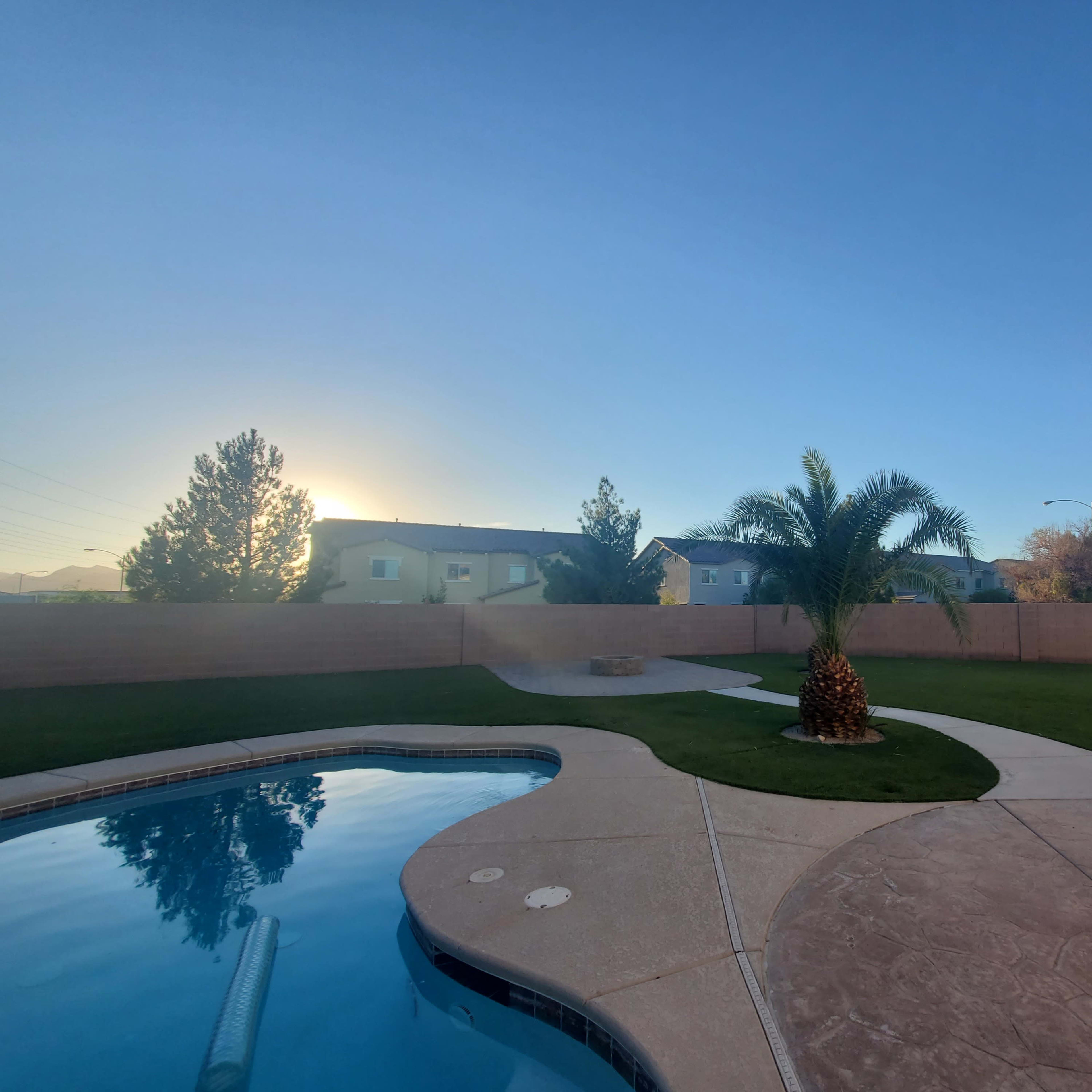 A clear blue sky overlooks a backyard with a swimming pool, palm tree, and well-maintained lawn surrounded by a low wall.
