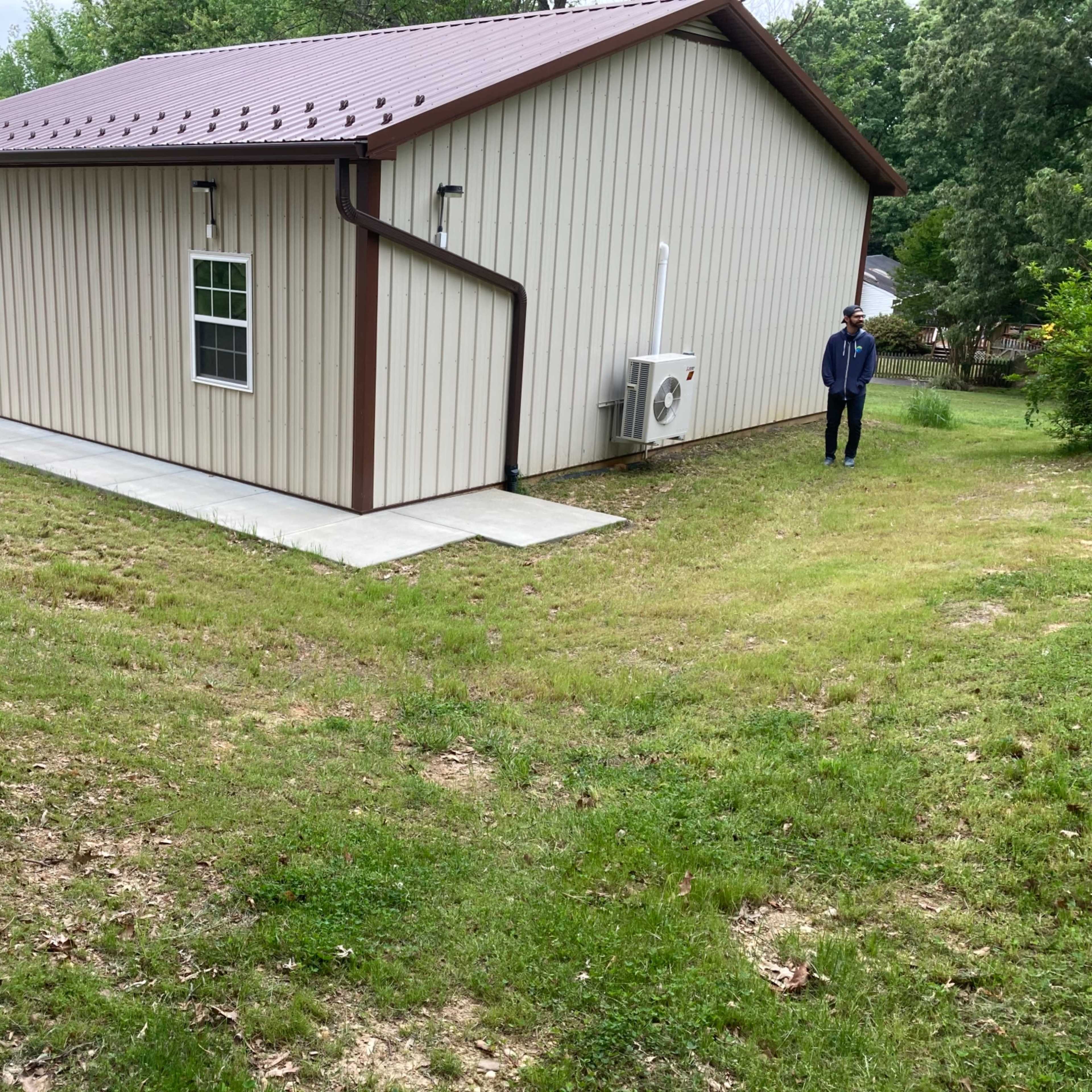 A person stands next to a beige metal building with a sloped lawn leading to a concrete pathway.